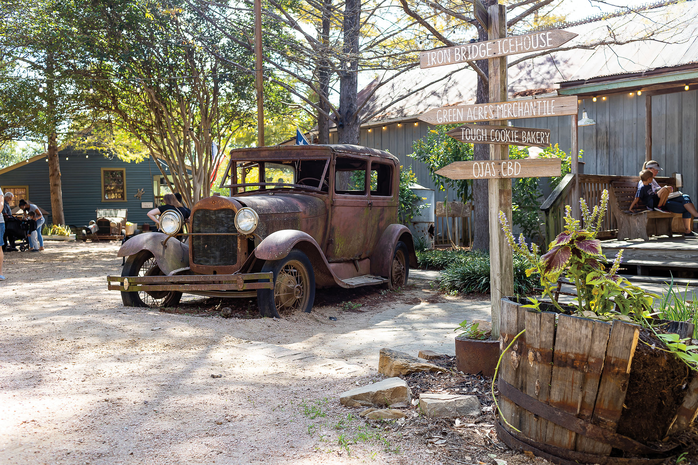 A rusted old-fashioned car sits in a gravel parking lot next to a wooden sign with arrows pointing different directions reading "Iron Bridge Icehouse," "Green Acres Mercantile," "Tough Cookie Bakery," and "Ojas CBD"