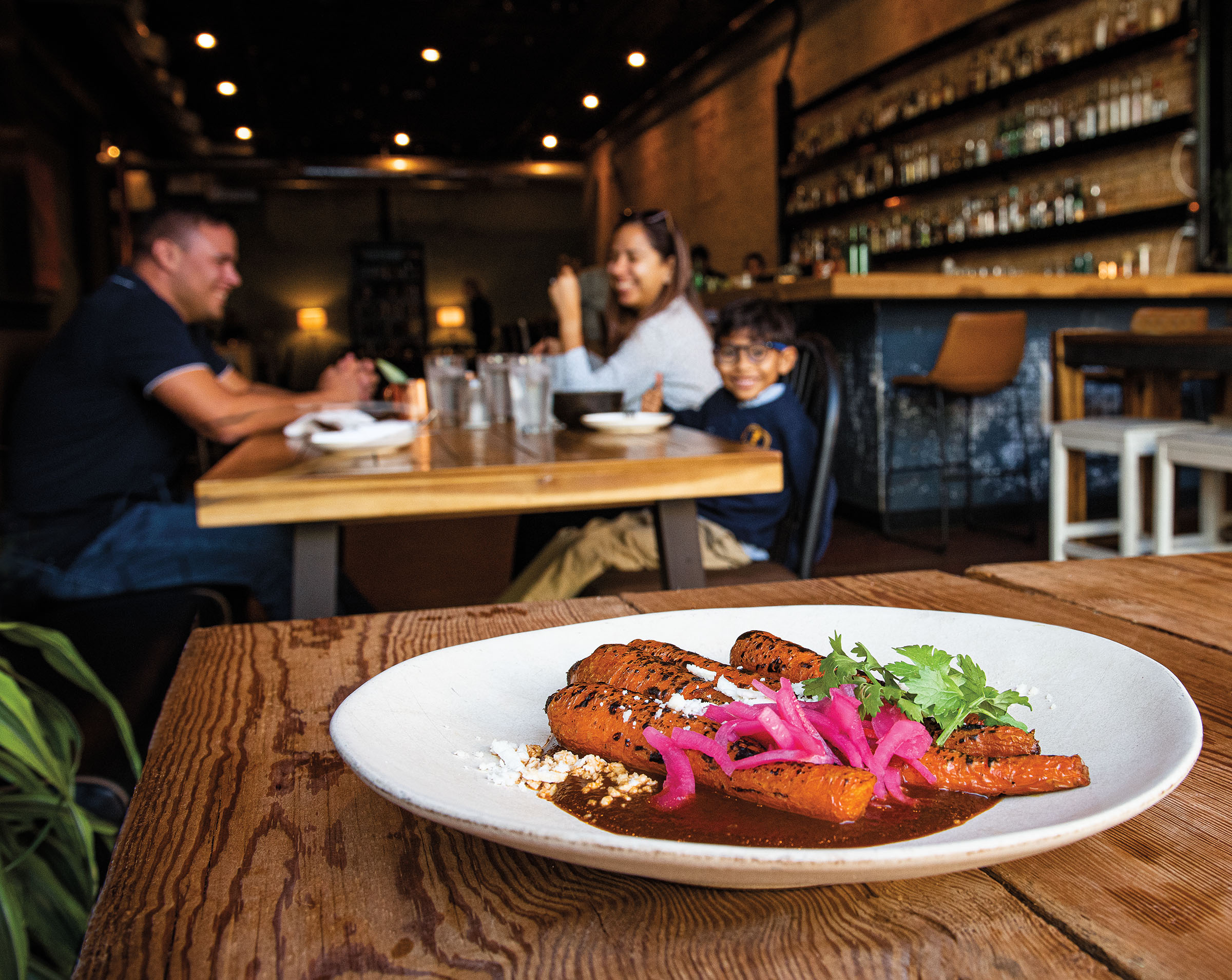 A wide white plate with carrots in a rich dark sauce topped with pickled red onions and cilantro. A table of three diners smiles in the background.