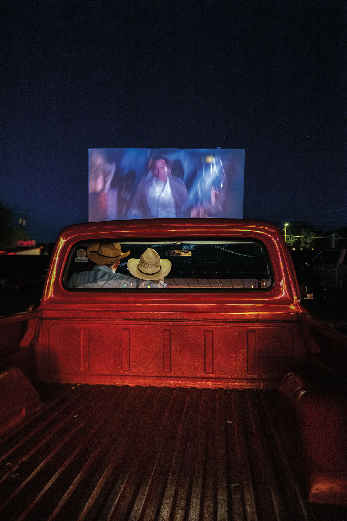 Man and woman in cowboy hats in the front seat of a red 1969 Ford F1100 pickup at a drive-in theater