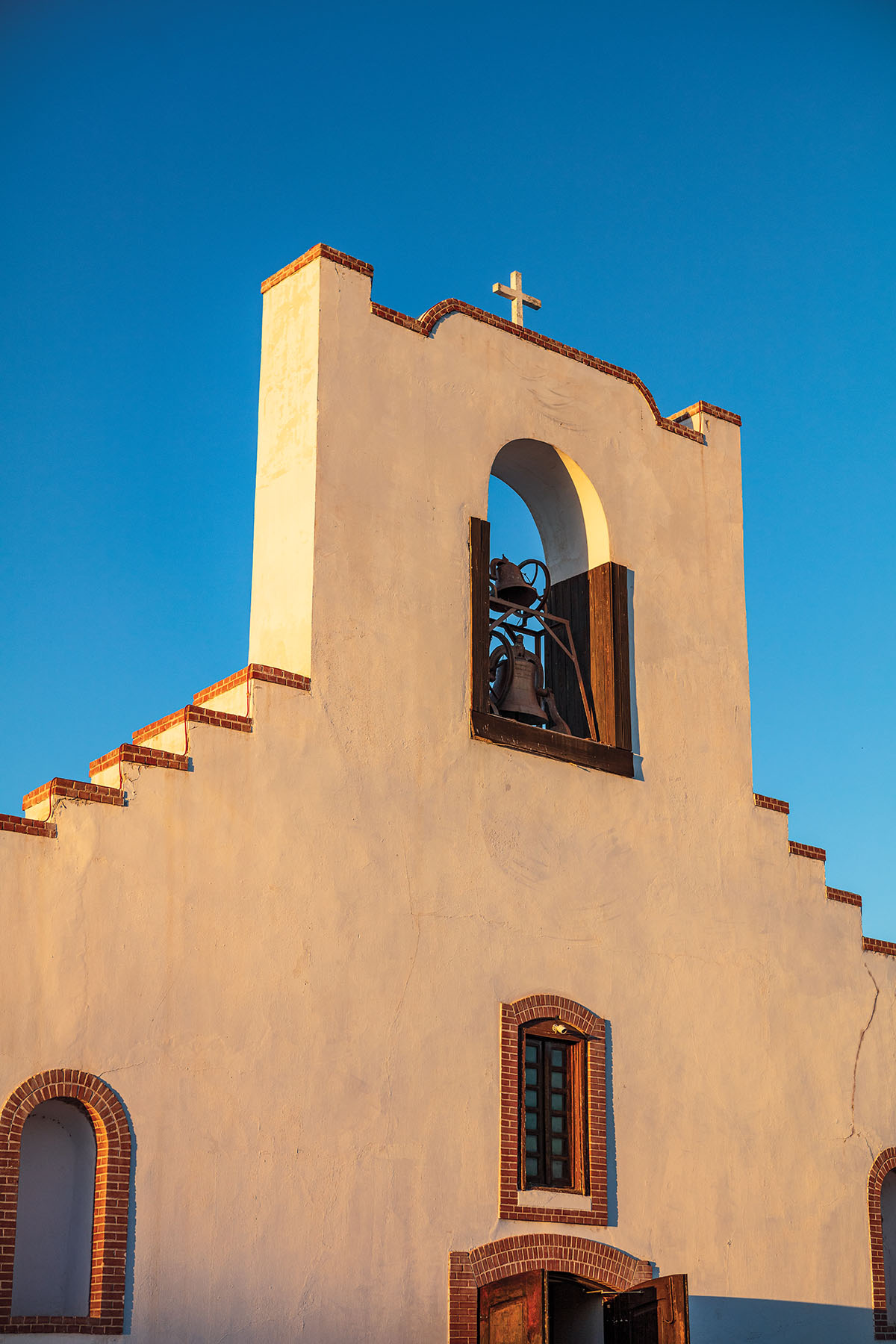 The outside of an adobe and brick church with a tall steeple and a bell inside