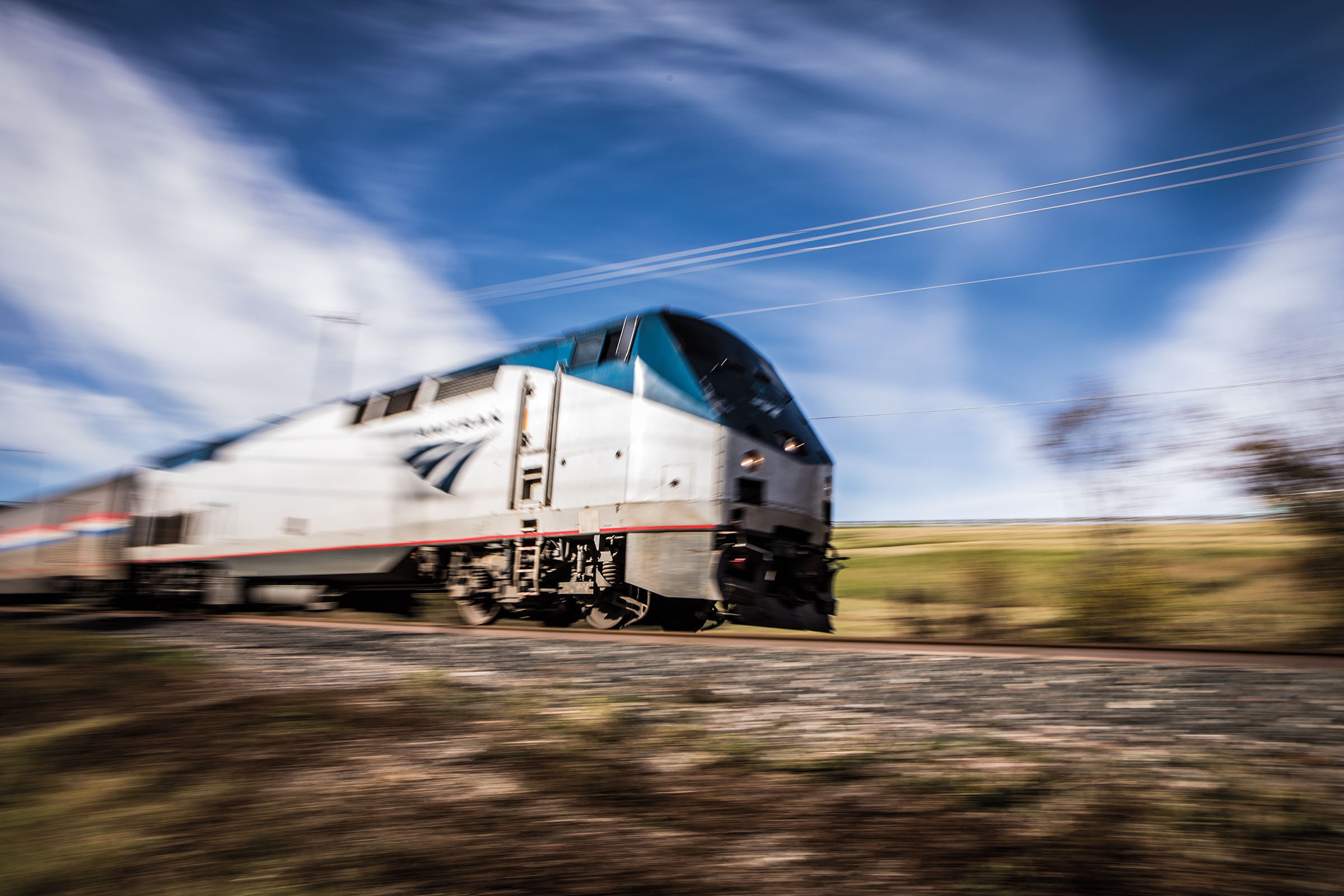 A blue and white locomotive whizzes past a landscape with a blue sky and gray train grade