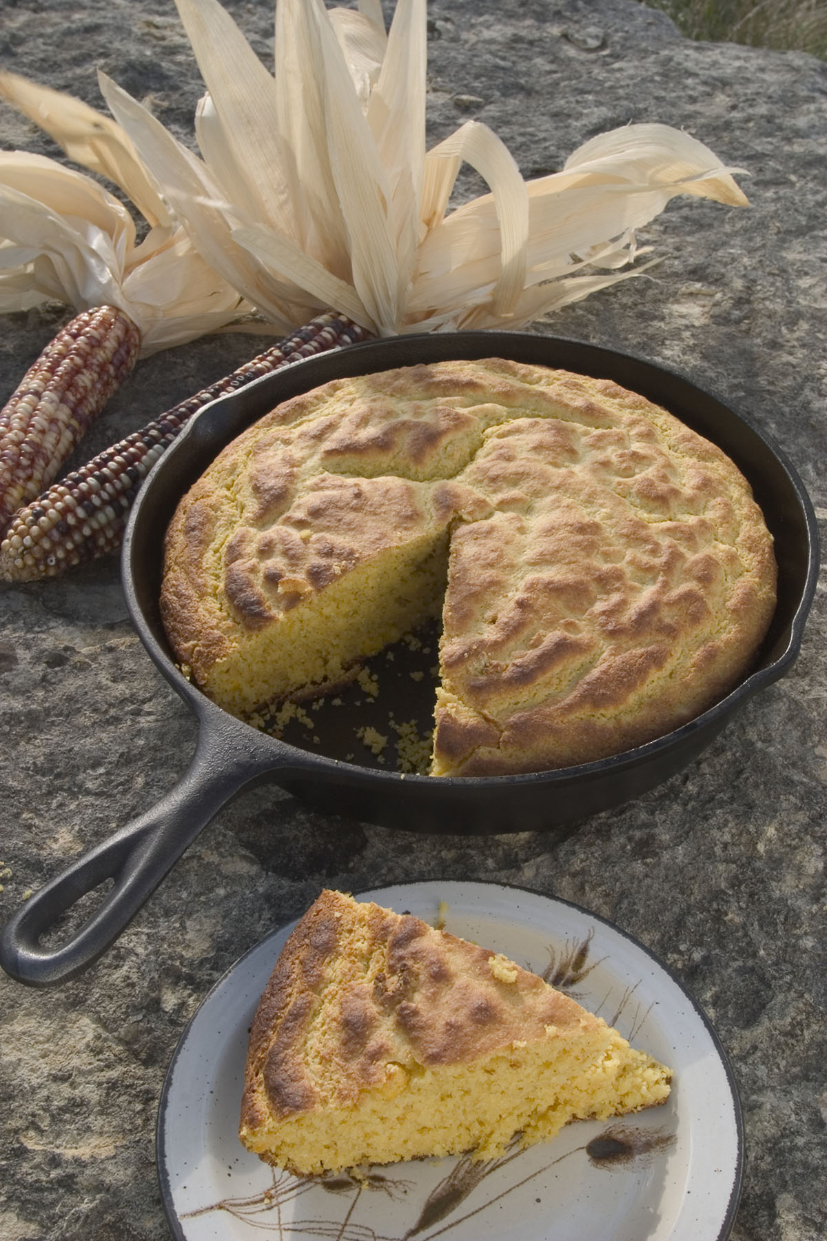 Golden cornbread with a slice missing from a large black cast iron skillet. Dried corn husks are visible just out of frame on a rocky background.