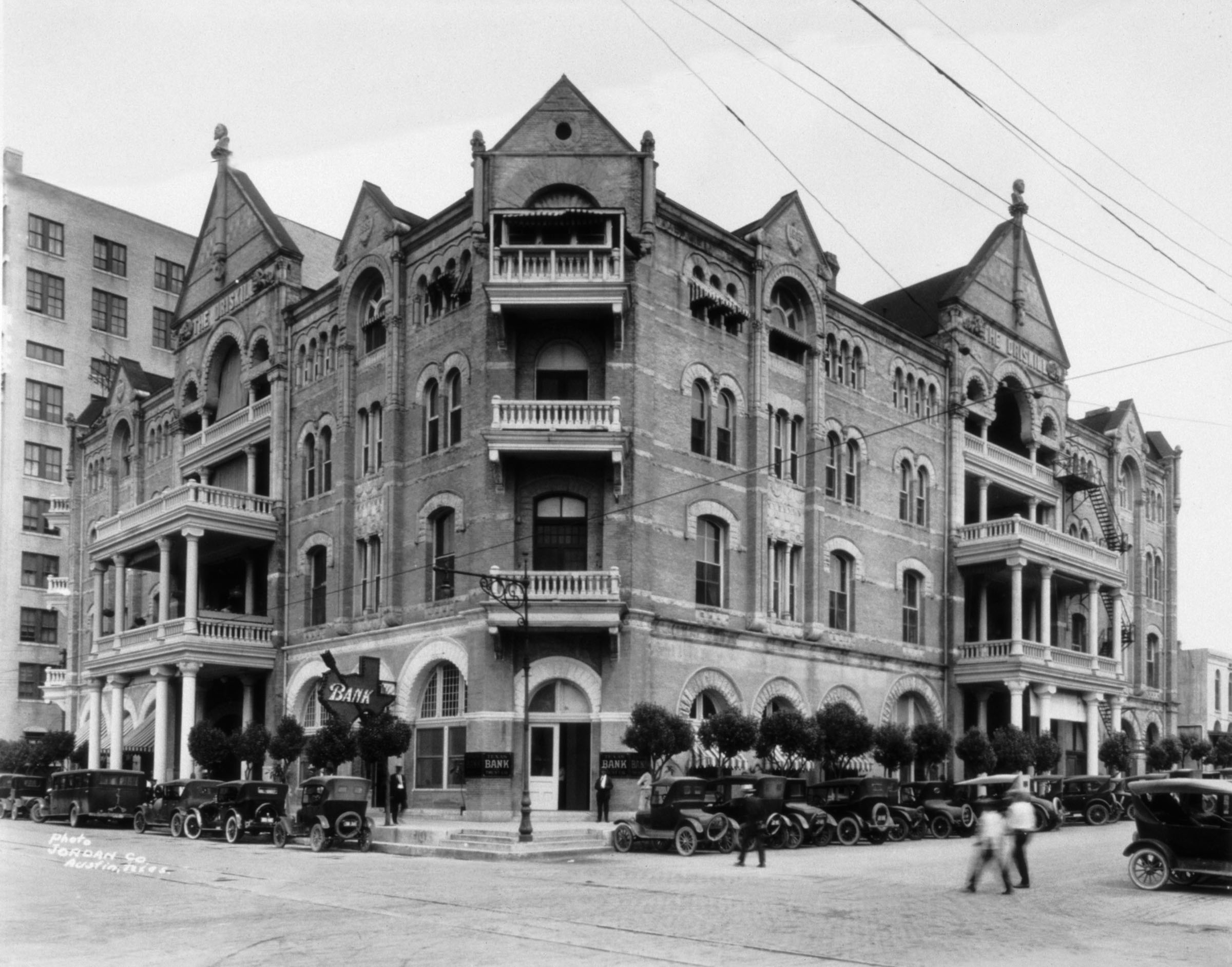 A black-and-white photo of the corner where the Driskill Hotel sits, with two people walking across the street and old cars parked along the curb.