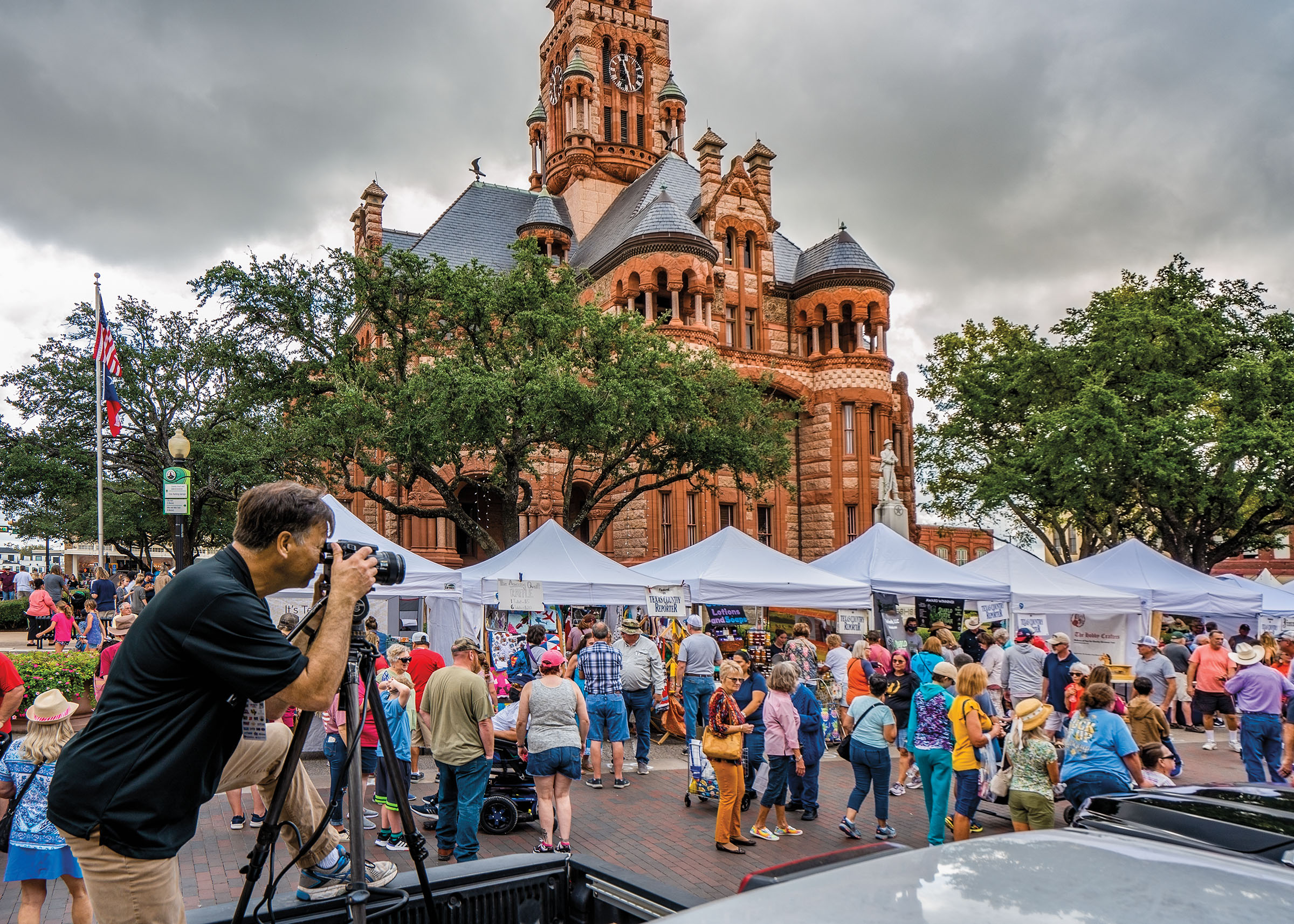 A man stands looking through a camera toward a crowd of people in front of white tents and a red-brick courthouse