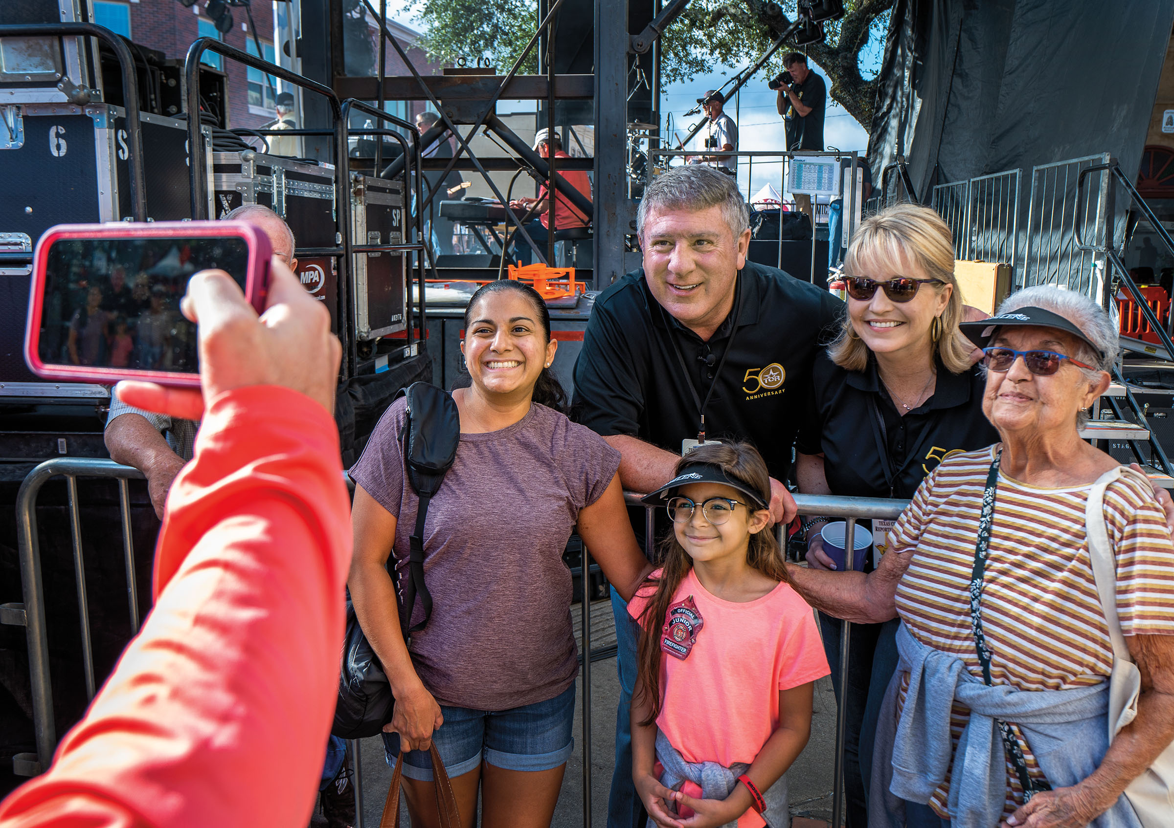 A group of people stand in front of a man and woman in black polo shirts and pose for a photograph. The setting appears to be a backstage area of a festival.