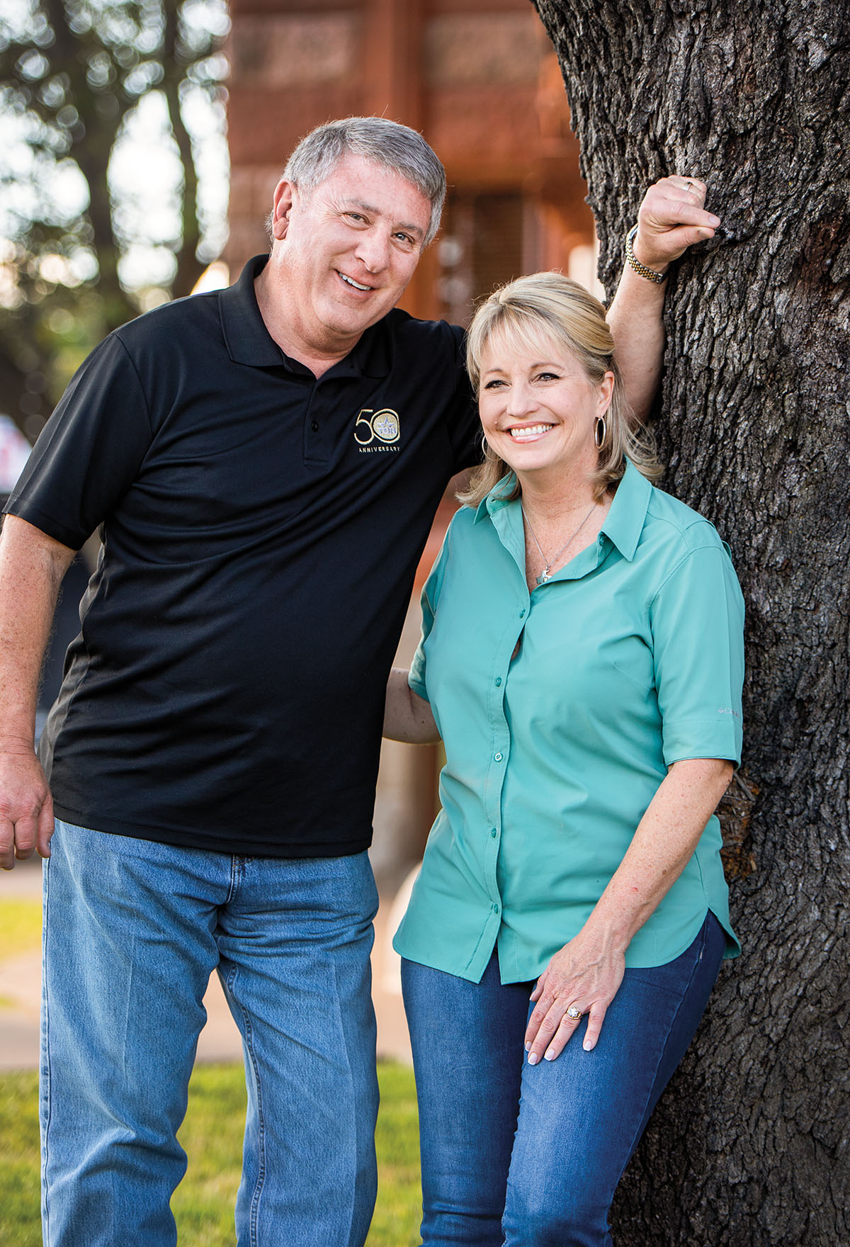 A man in a black polo shirt stands next to a woman in a green button-down shirt leaning on a large tree
