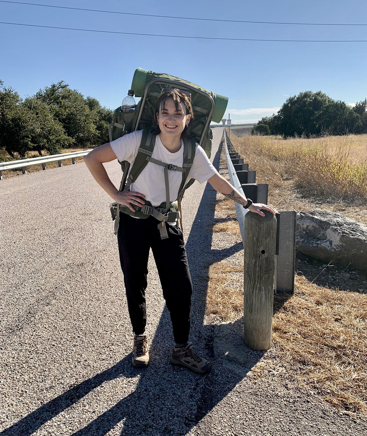 A woman in a t-shirt and green backpacking pack stands at the side of a road under blue sky