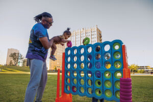 A man holds a young child up so she can place a marker in a larage Connect 4 set, in a park with green grass and a tall building in the background