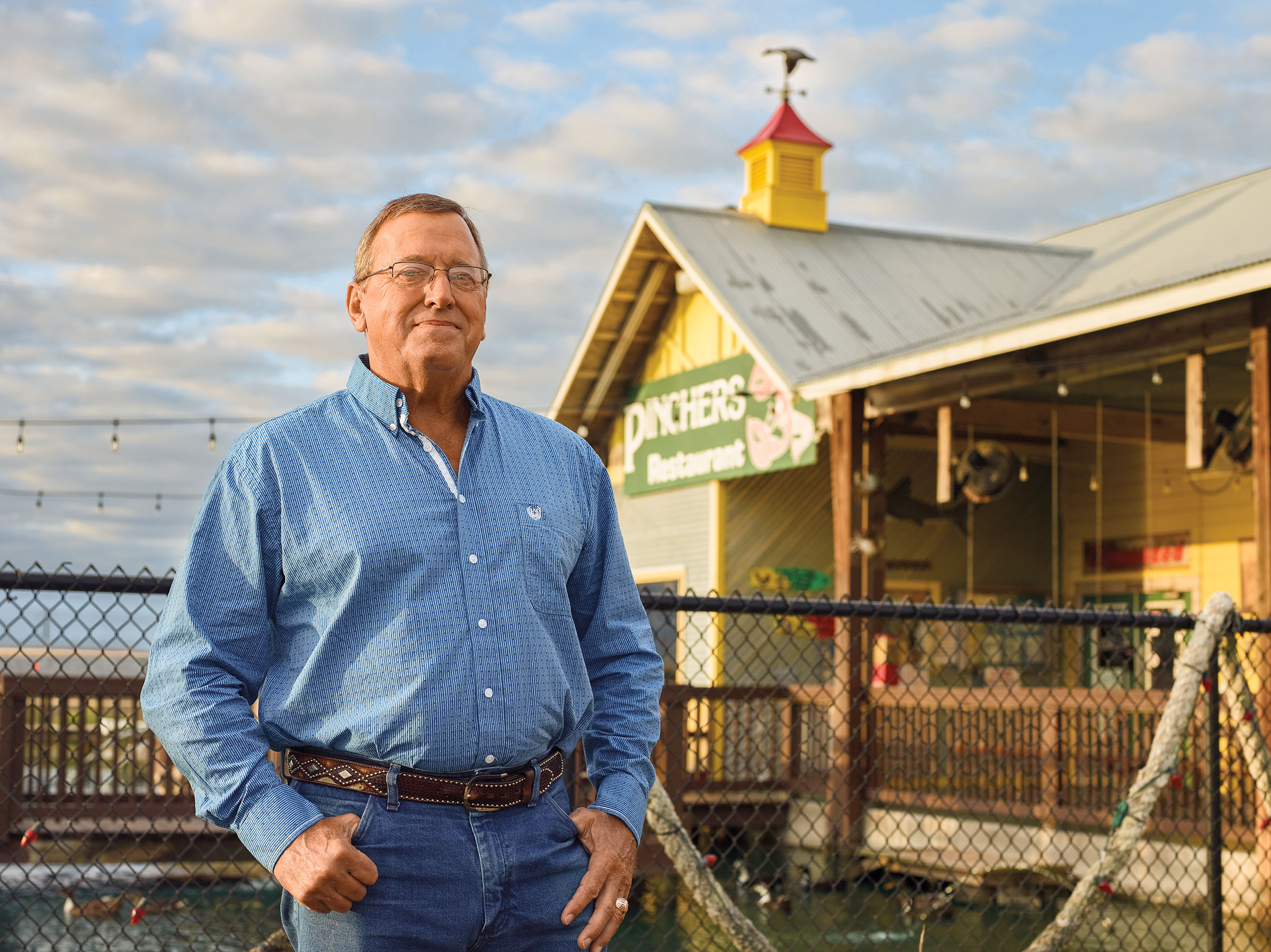 A man in a blue button-down shirt and blue jeans stands in front of a yellow building with a sign reading "Pinchers"