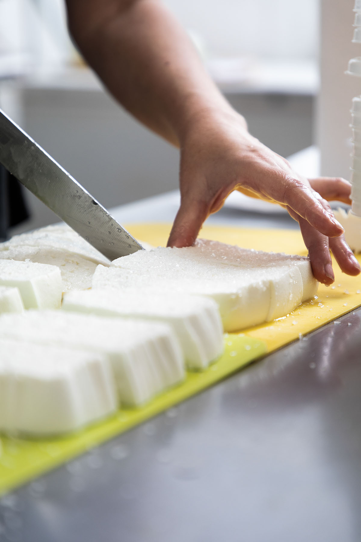 A person holds large white curds of cheese and cuts them on a yellow cutting board
