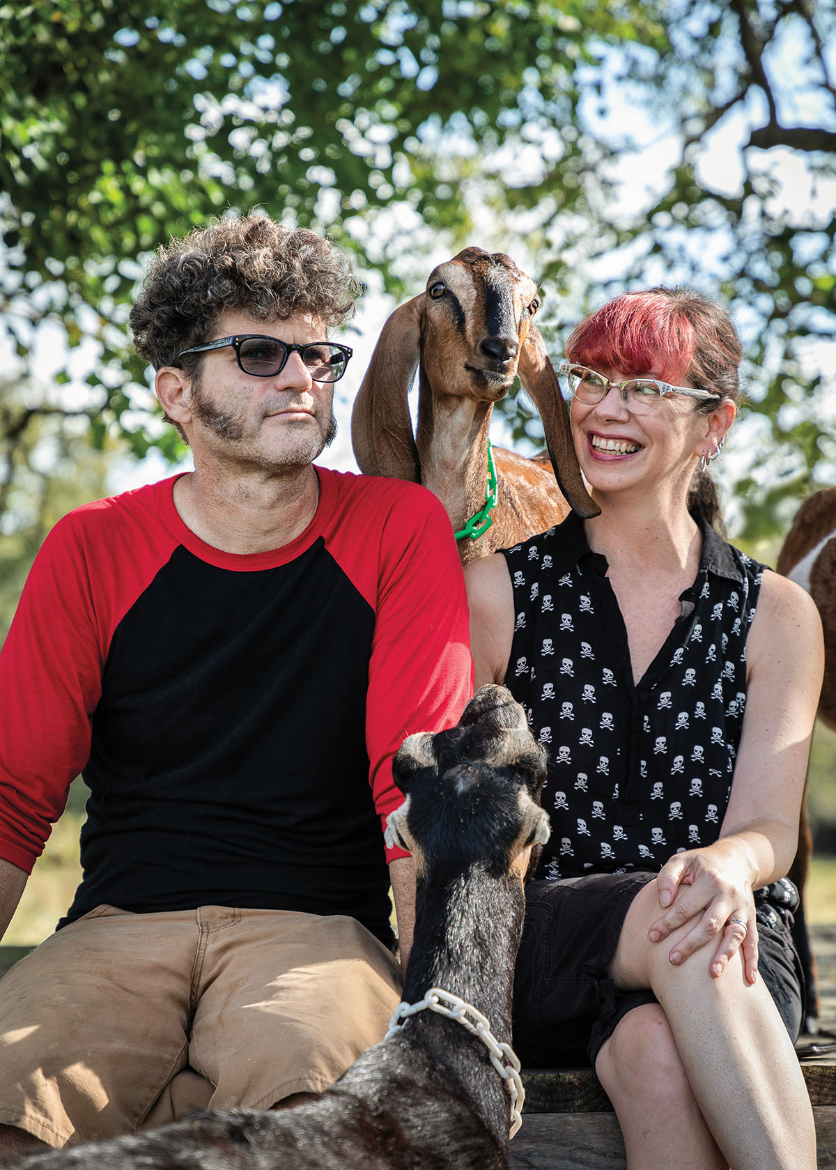 Two people sit smiling with a goat between them in a green outdoor setting