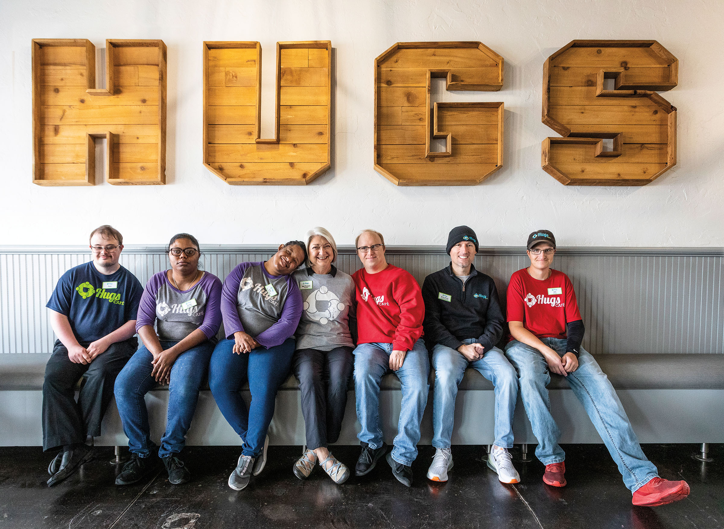 A group of people in t-shirts and jeans sit in front of a large wooden sign reading "HUGS"