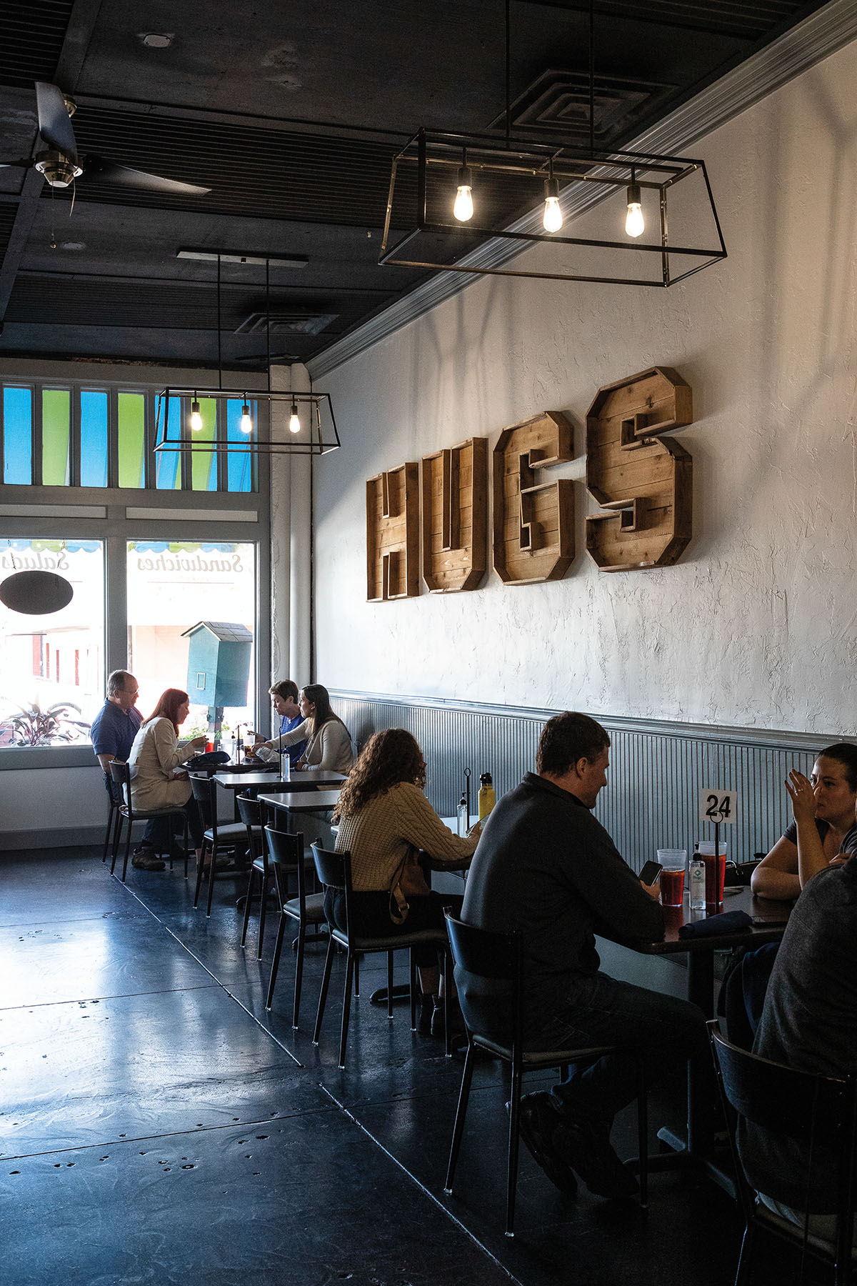 The white and beadboard wall of the restaurant with a wooden sign reading "HUGS" above several tables of diners