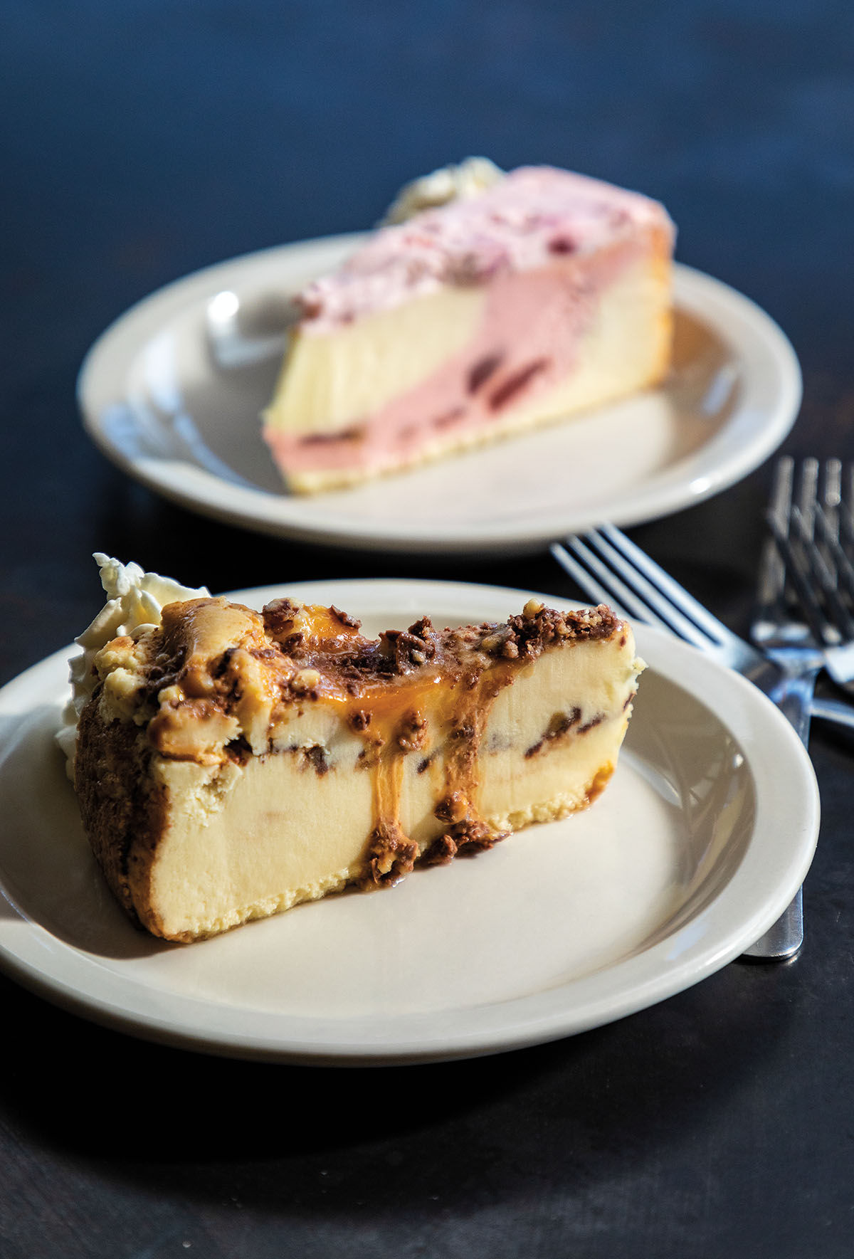 In the background, a piece of cheesecake with pink swirls and fruits. In front, a cheesecake with a caramel, nut and chocolate topping
