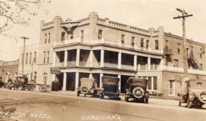 A sepia photograph of a three-story stone hotel with several vintage cars parked out front