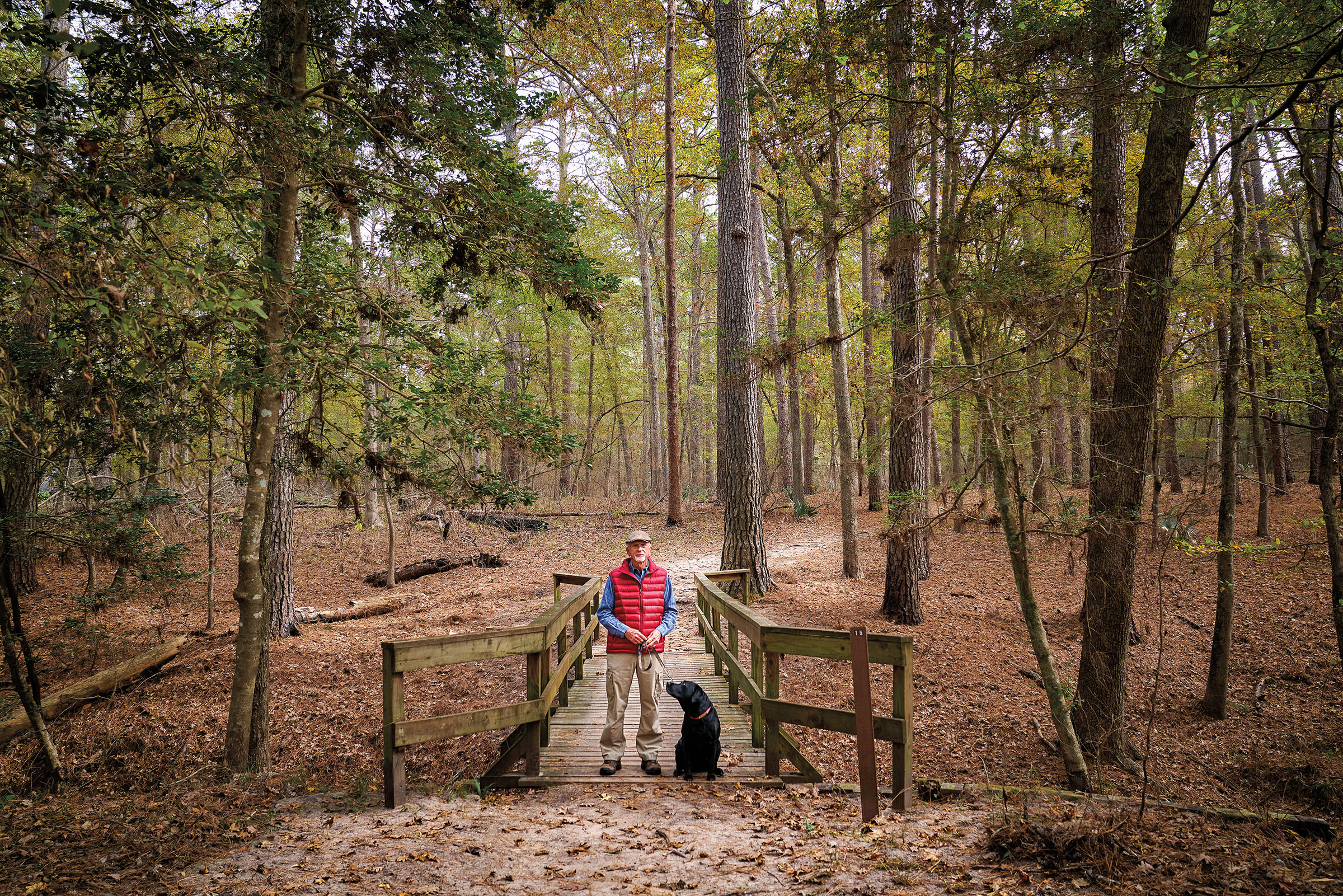 A man in a red vest stands in the middle of a park next to a black lab