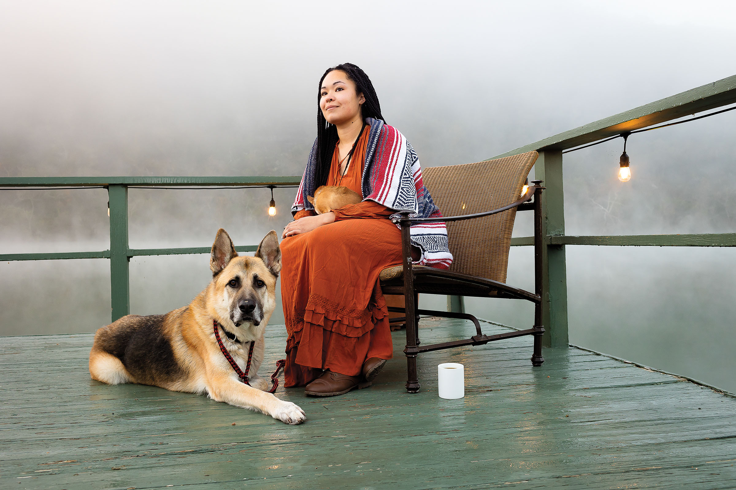 A woman in a long orange skirt and blanket sits in a chair next to her dog in foggy lighting