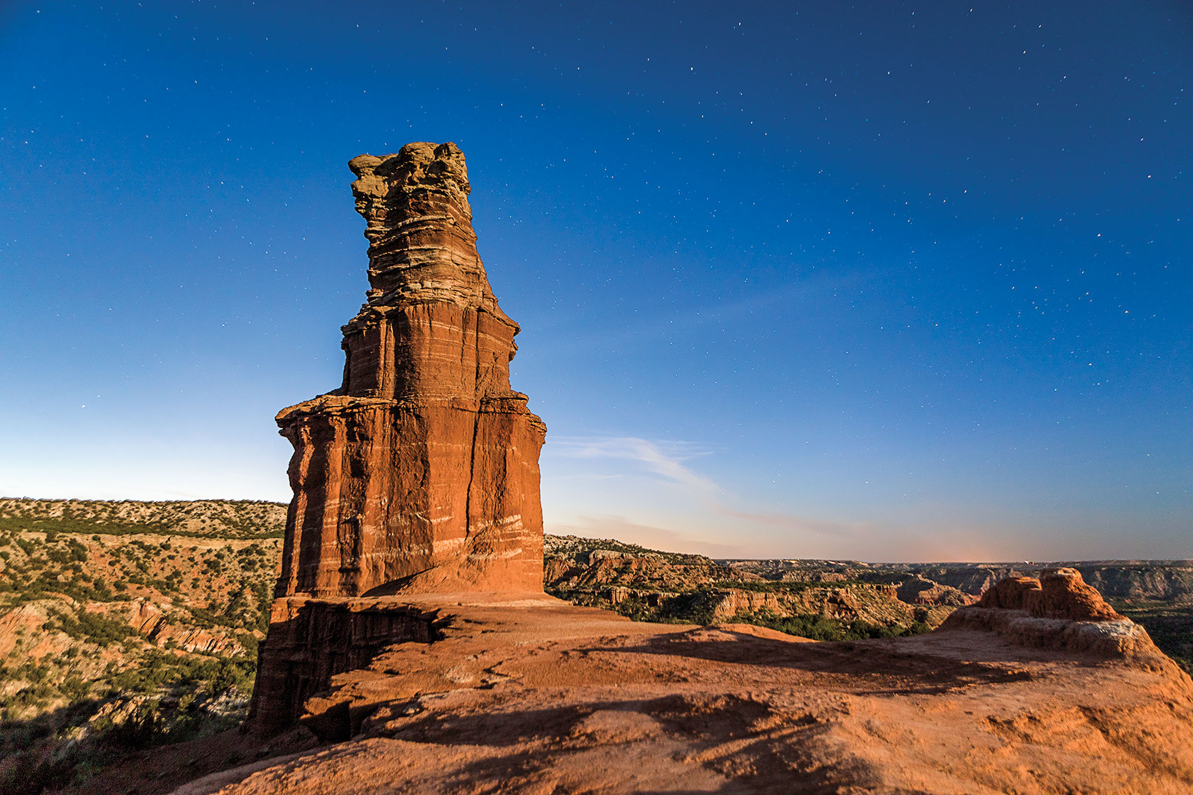 A tall, golden-brown rock formation amid a desert landscape under deep blue sky