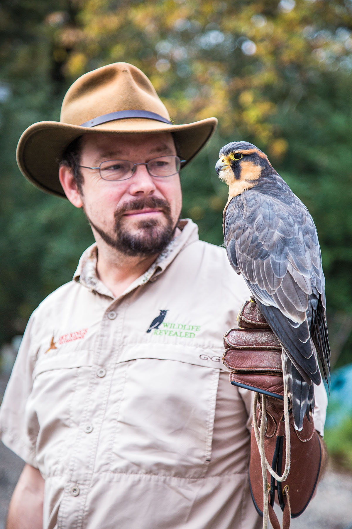 A man in a wide-brimmed brown hat stands with a bird perched on his gloved hand