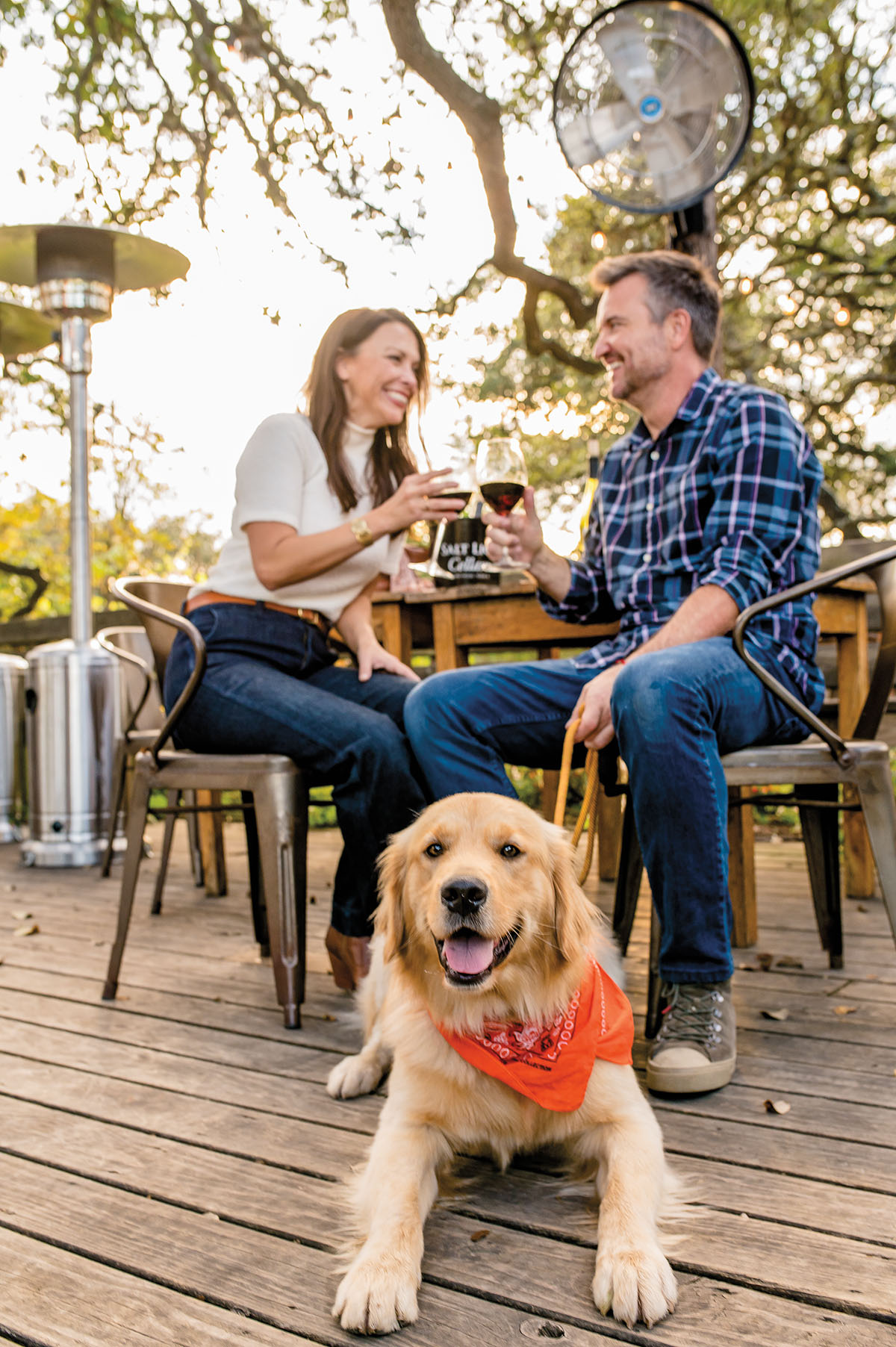 A golden retriever wearing an orange bandana looks at the camera while two people laugh and sip wine in the background