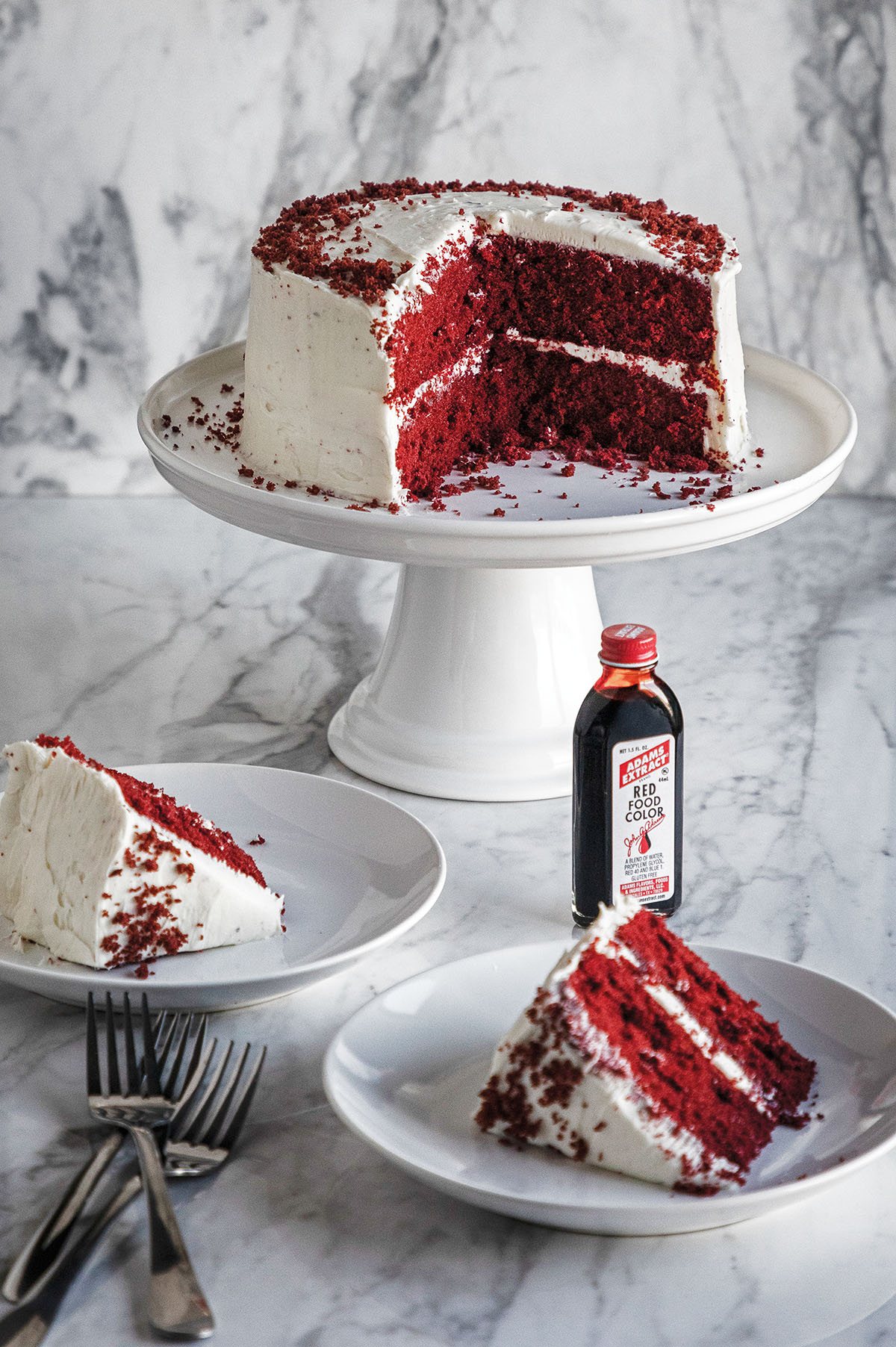 A white-frosted red velvet cake sitting on a white platter next to a bottle of Adam's Extract