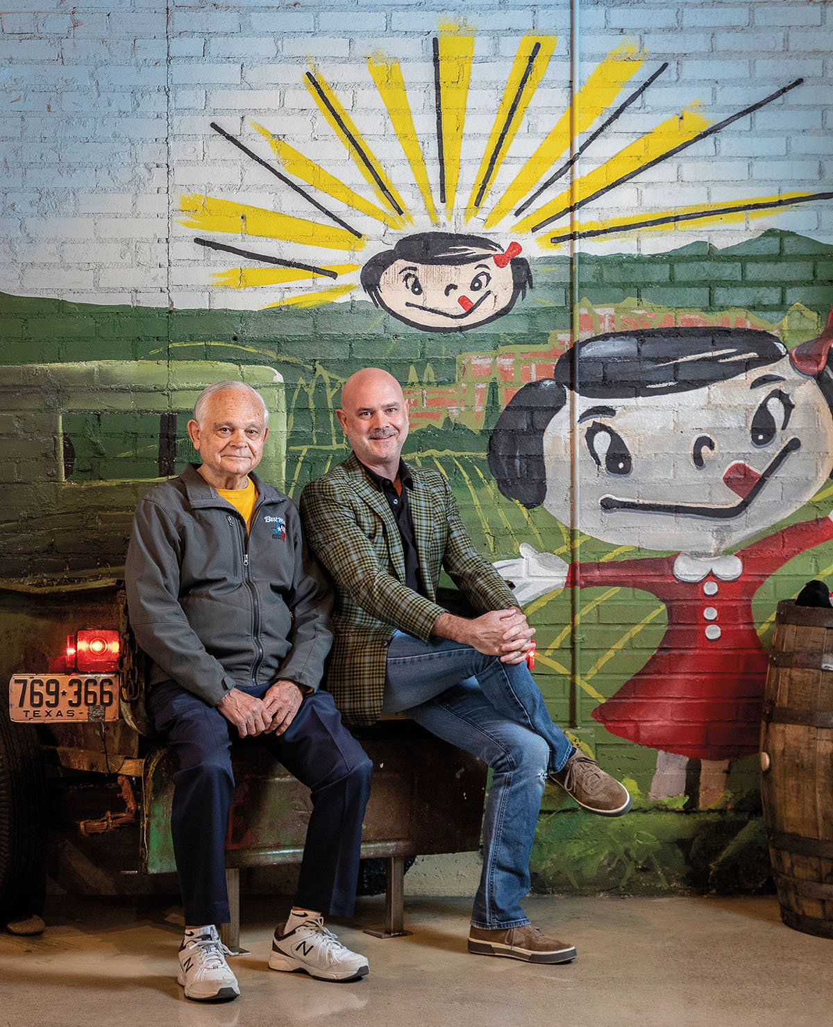 Two men sit on a small bench in front of a mural painted with the Best Maid cartoon mascot and a sun