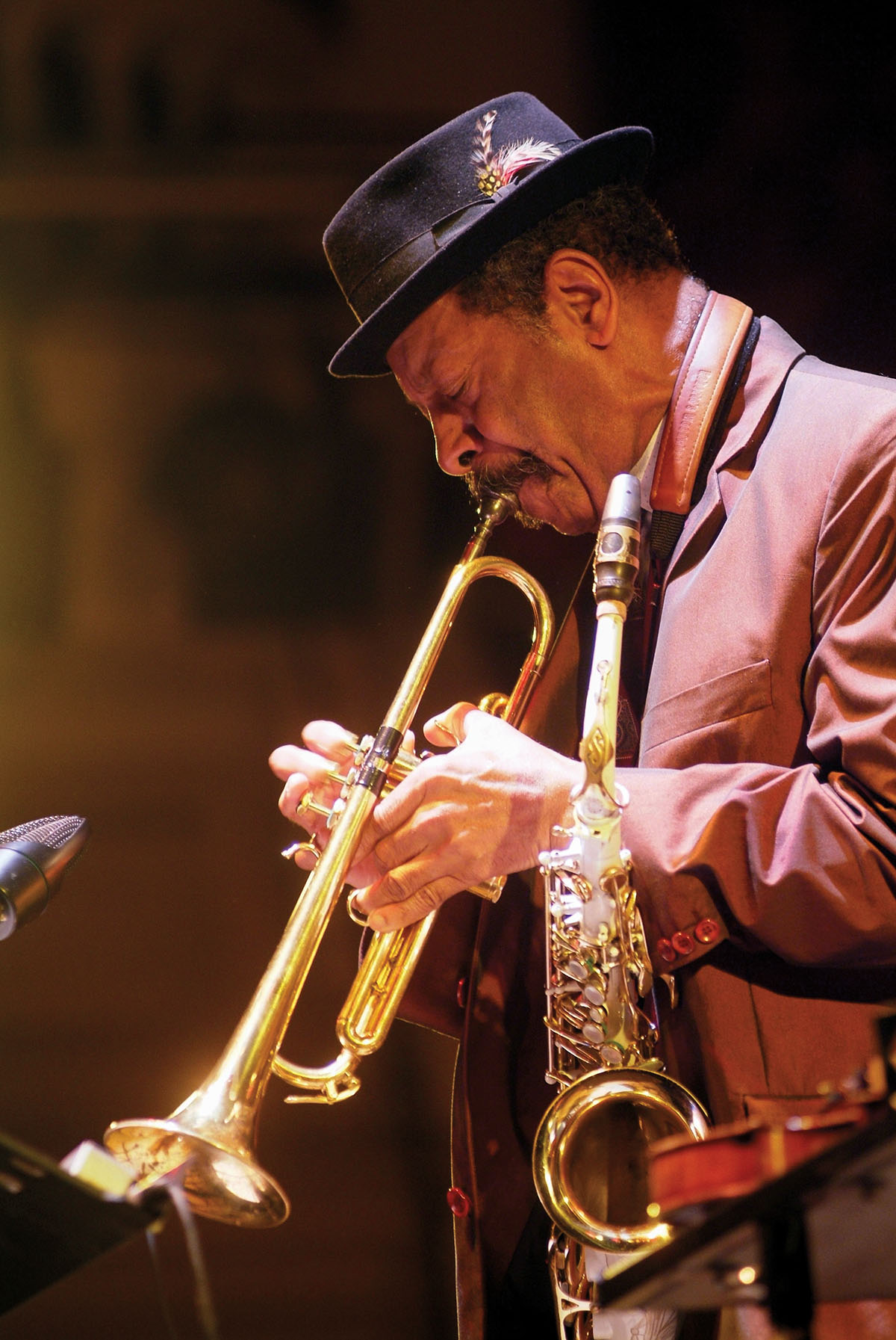 A man in a small black hat and suit jacket plays a golden trumpet