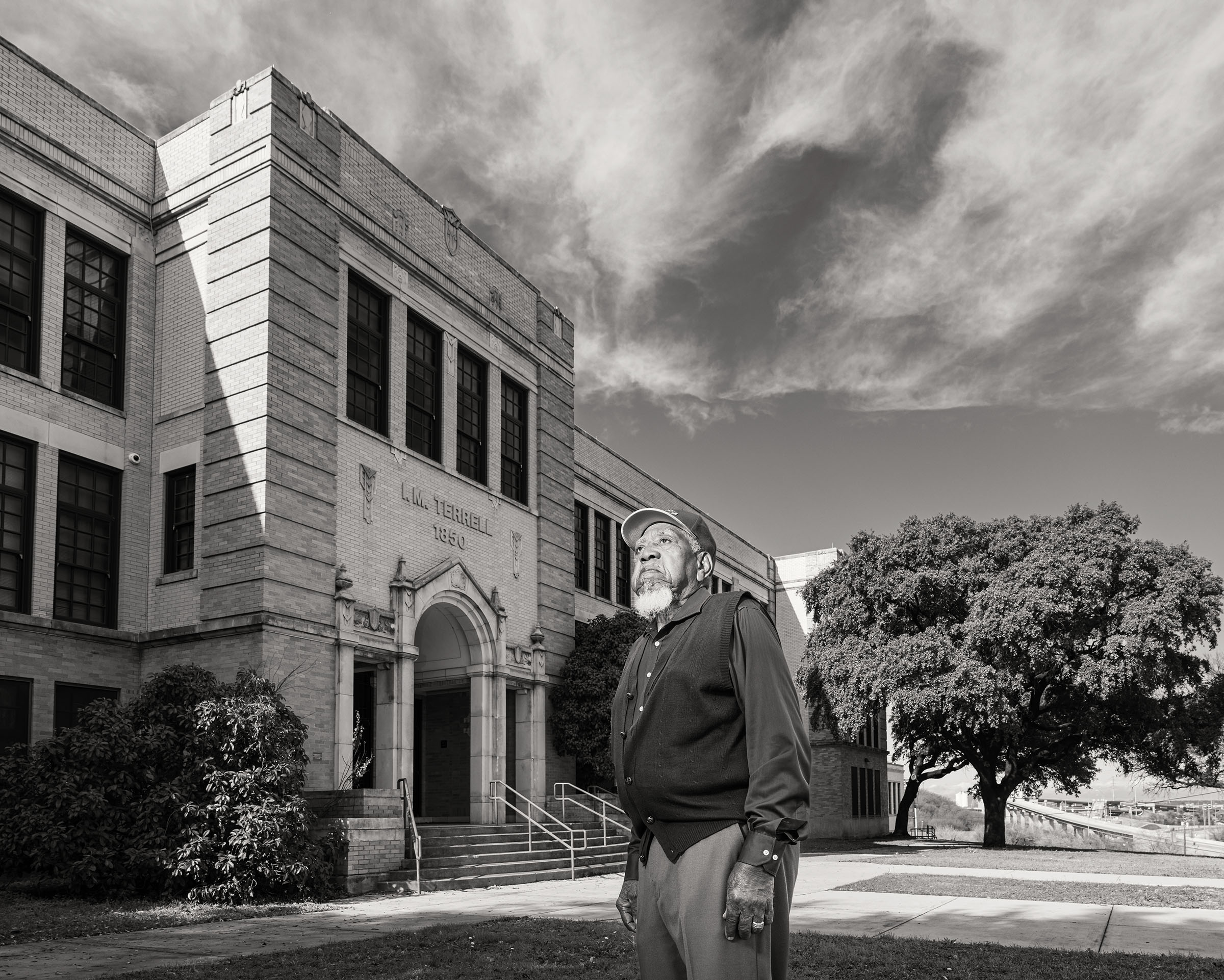 An older man in a vest and long-sleeved shirt walks in front of a brick school in this black and white photograph