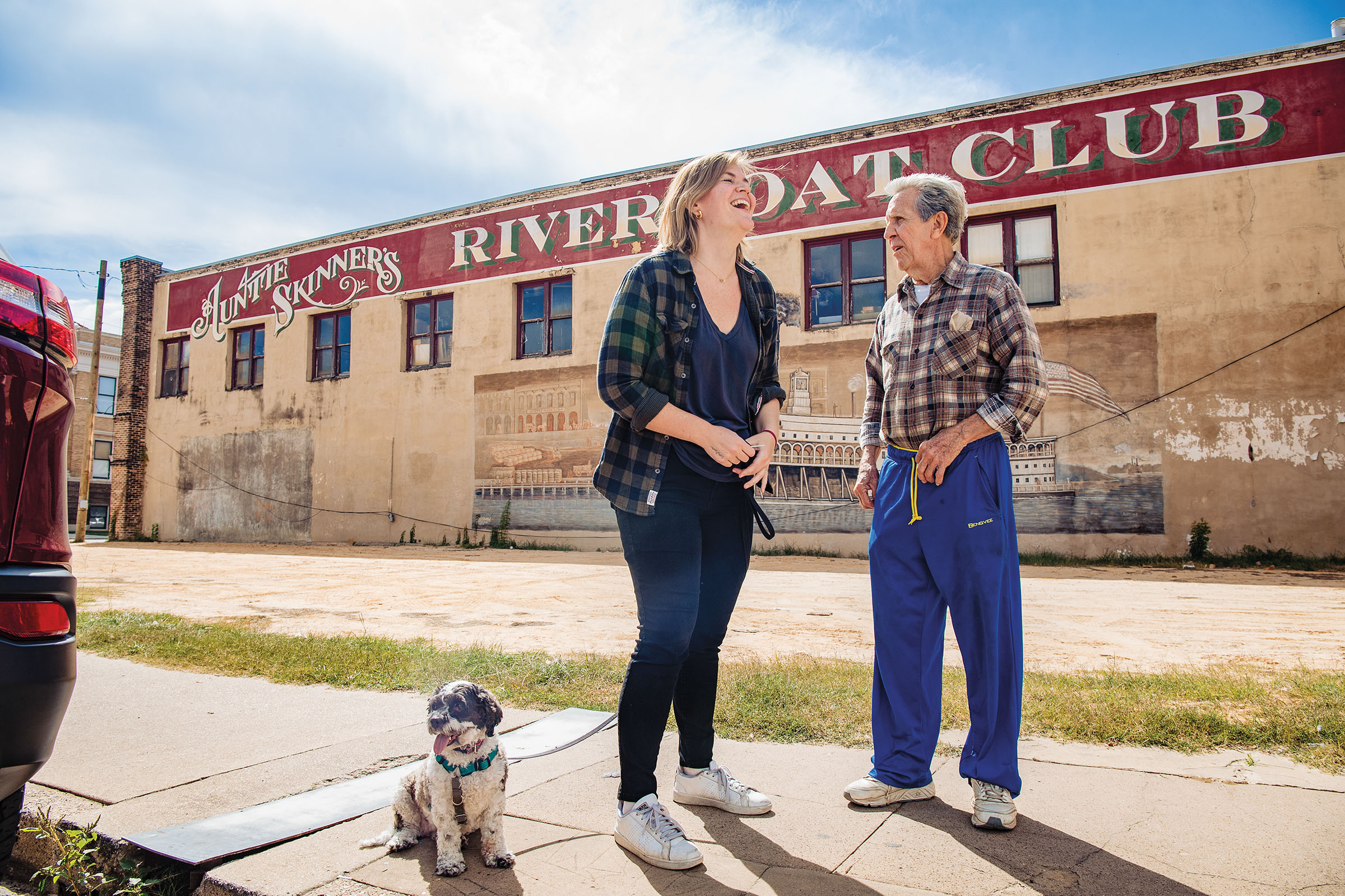 The author and her dog stand and laugh next to an older gentleman wearing jeans standing in front of an old building