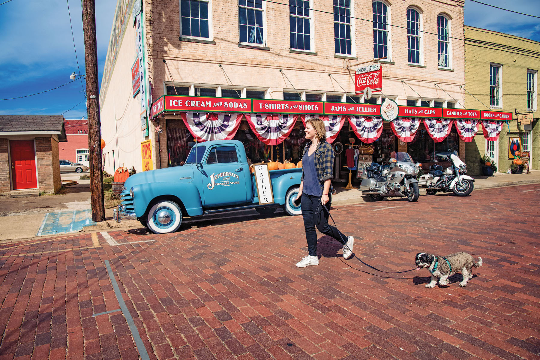 A woman walks her small dog down a brick street in front of a brick building decorated with red, white, and blue bunting