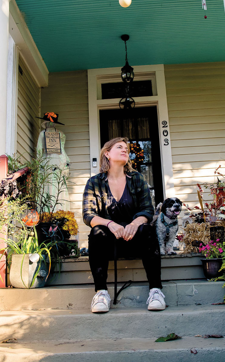 A woman and a small dog sit on the steps of an ornately decorated house with potted plants and teal painted ceiling