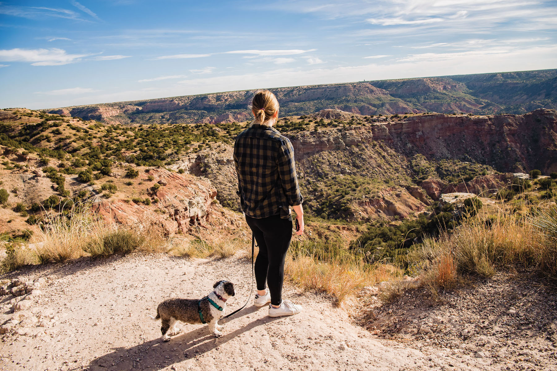 A woman stands with her dog on a dirt path looking at the brown-green canyons of Palo Duro