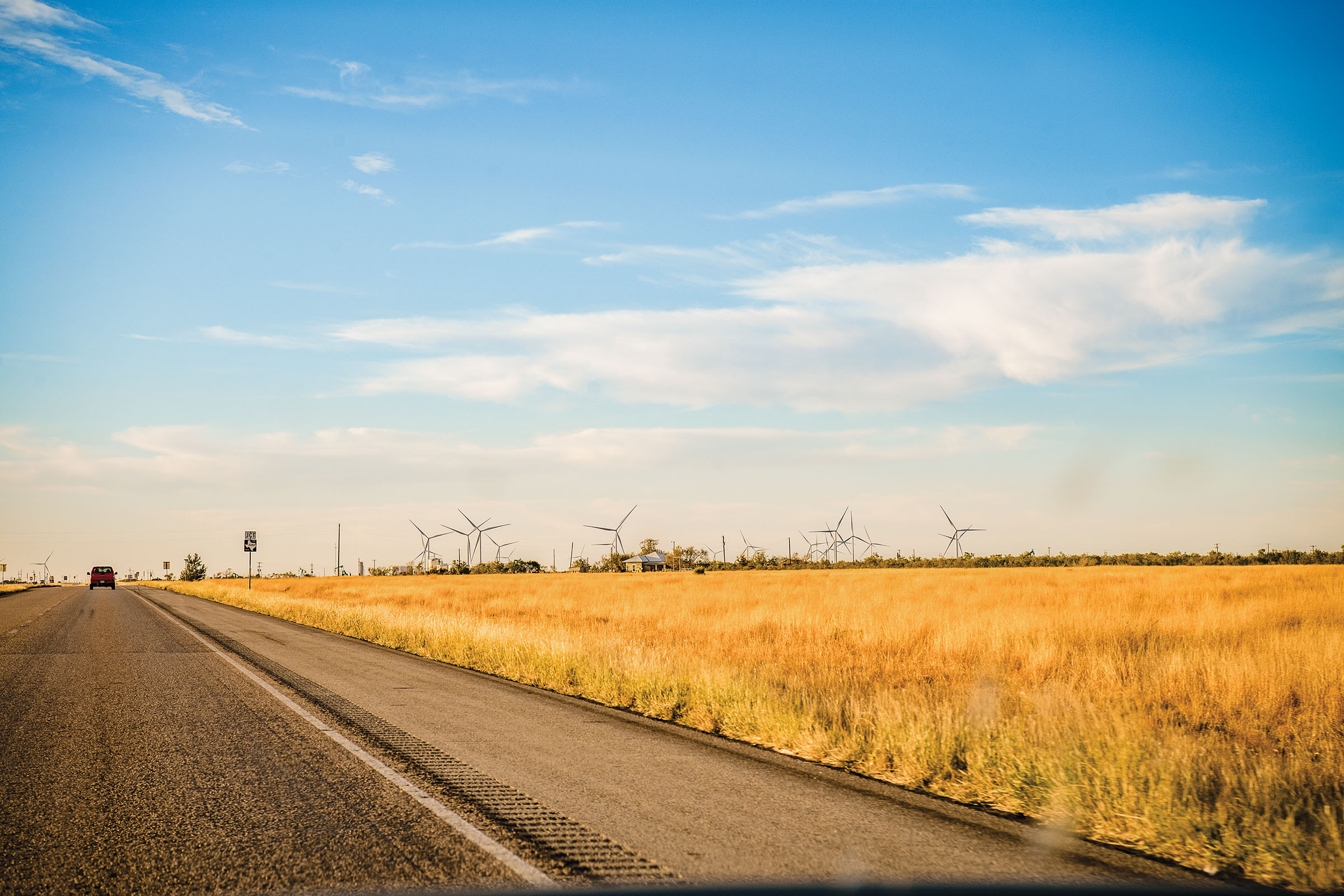 Tan grasslands next to the shoulder of a black highway under a blue sky with a few clouds