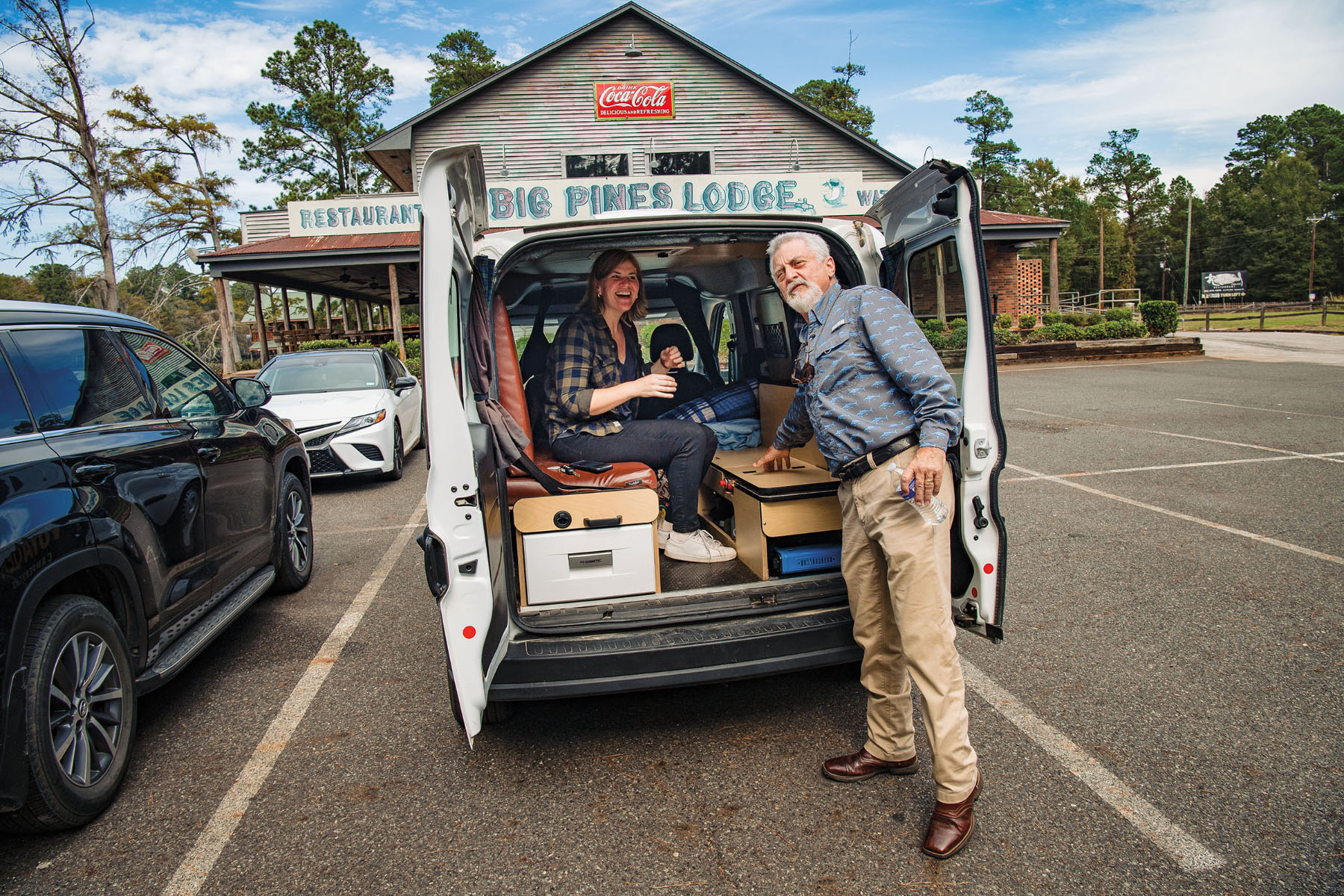 A woman sits and laughs inside a van while a man stands outside leaning toward the van in a parking lot