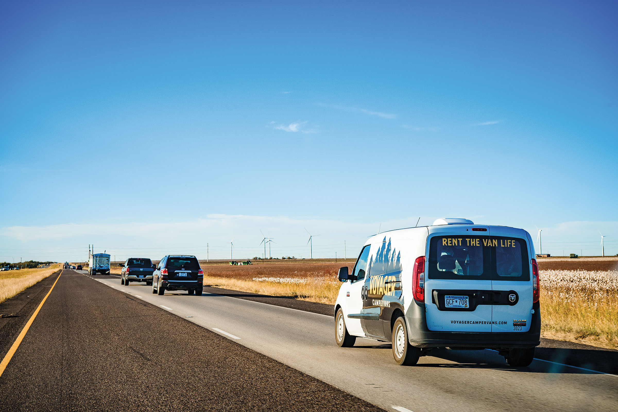 A small white van travels under a bright blue sky with tan fields and a highway with several cars