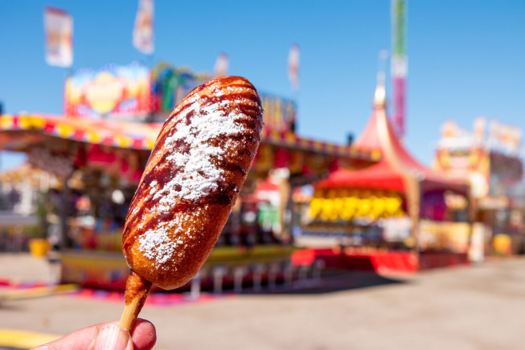A person holds a stick of a golden brown dessert covered with chocolate sauce and powdered sugar. Blurred in the background is a colorful, carnival-like scene