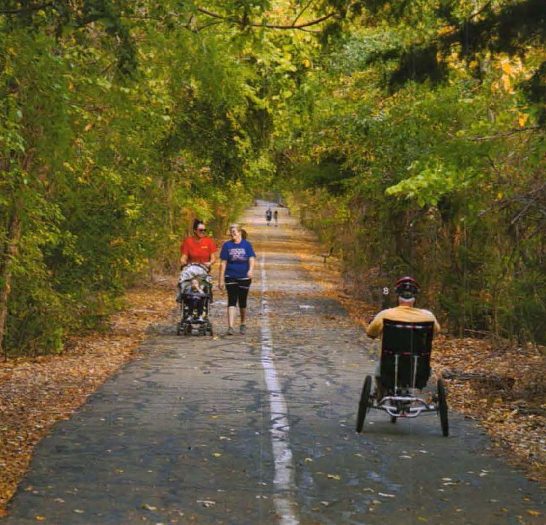 A man in a three-wheeled vehicle pedals next to two women walking with a stroller