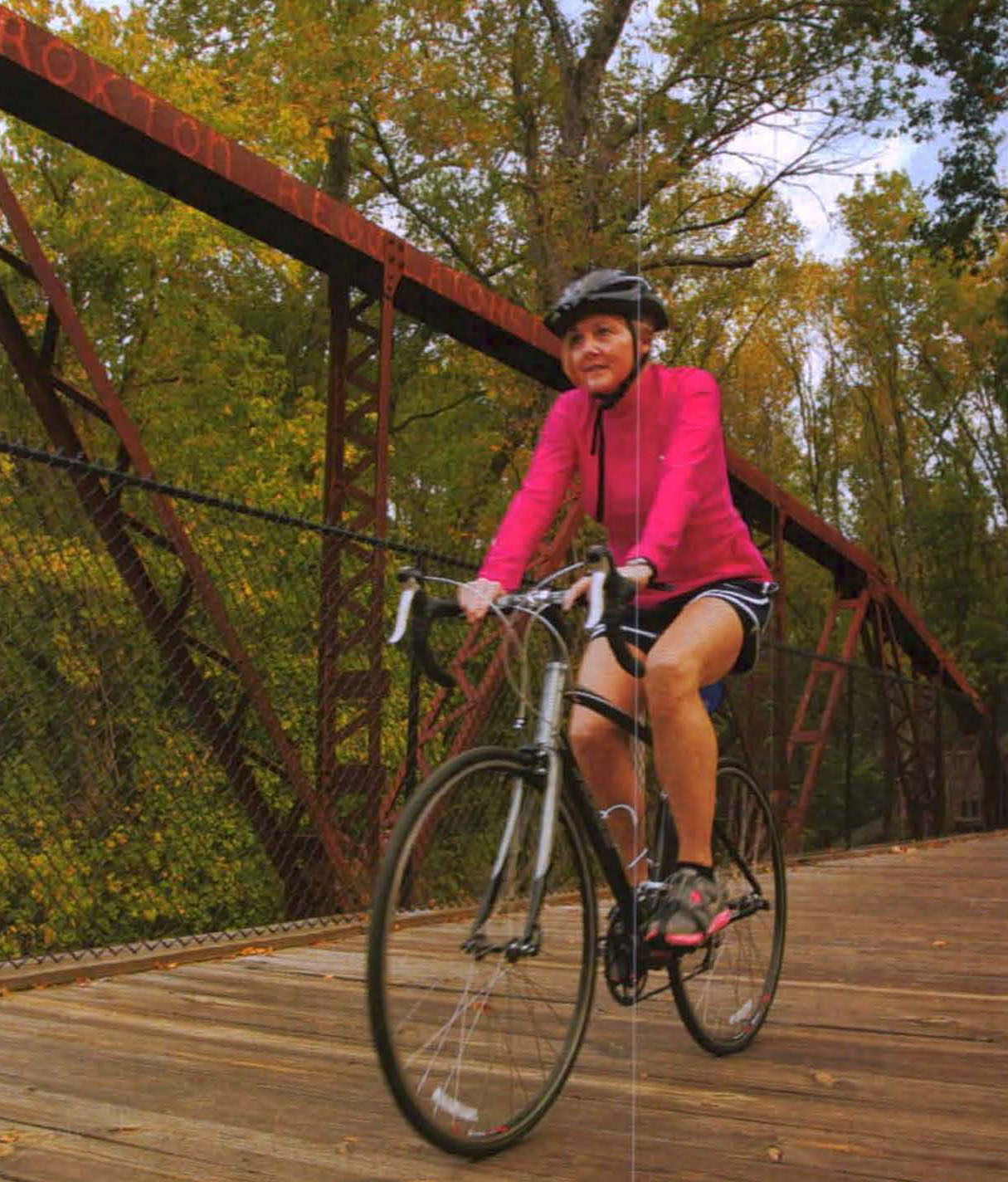 A woman in a pink jacket rides a bike across a wood-based bridge