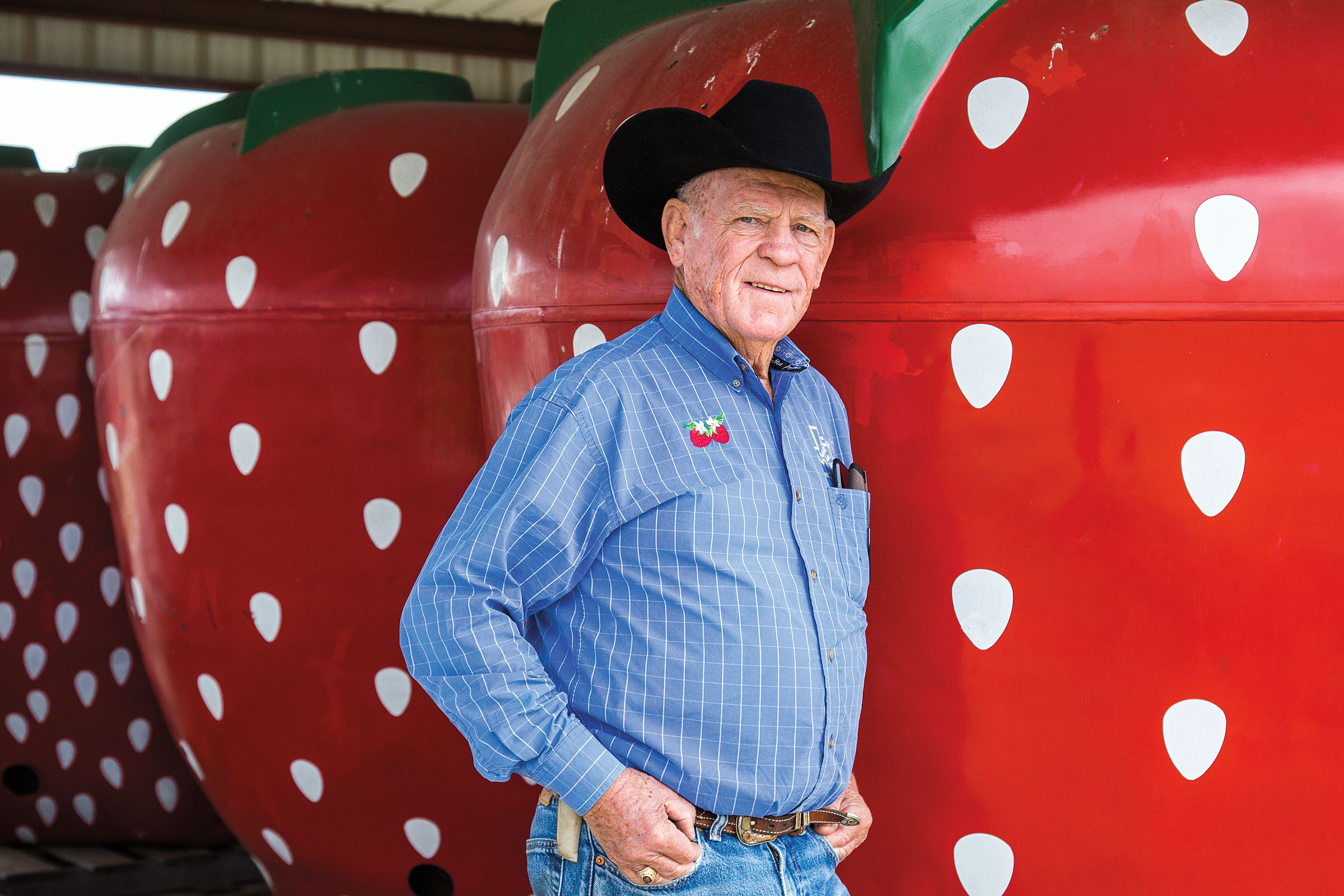 A man in a black cowboy hat stands in front of a large painted strawberry with white seeds