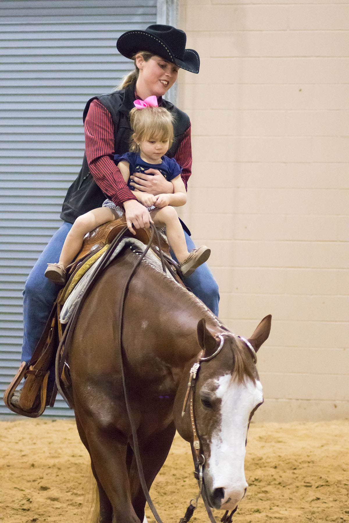A woman in a black cowboy hat holds a young child on a brown horse
