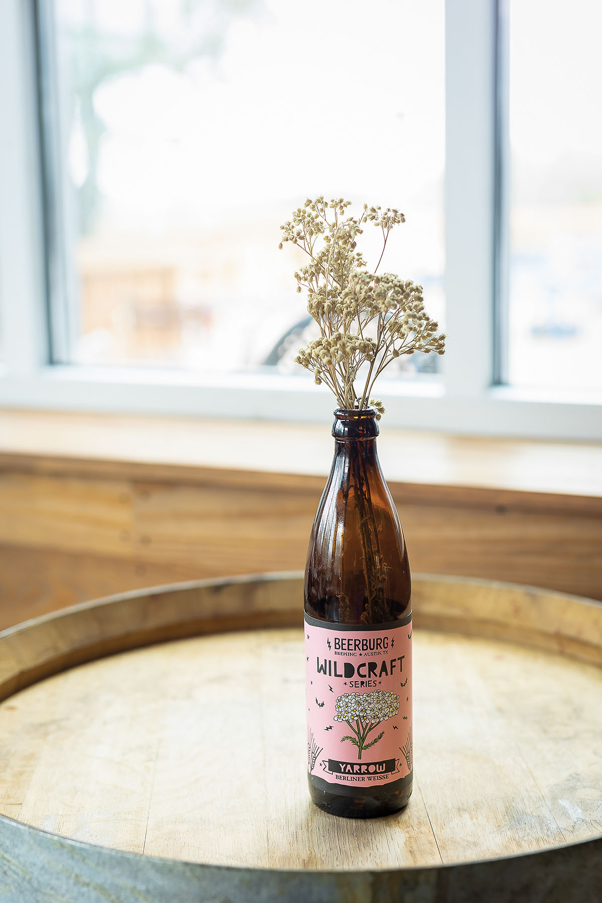 A small cut flower sticks out of the top of a pink-labeled beer bottle reading 