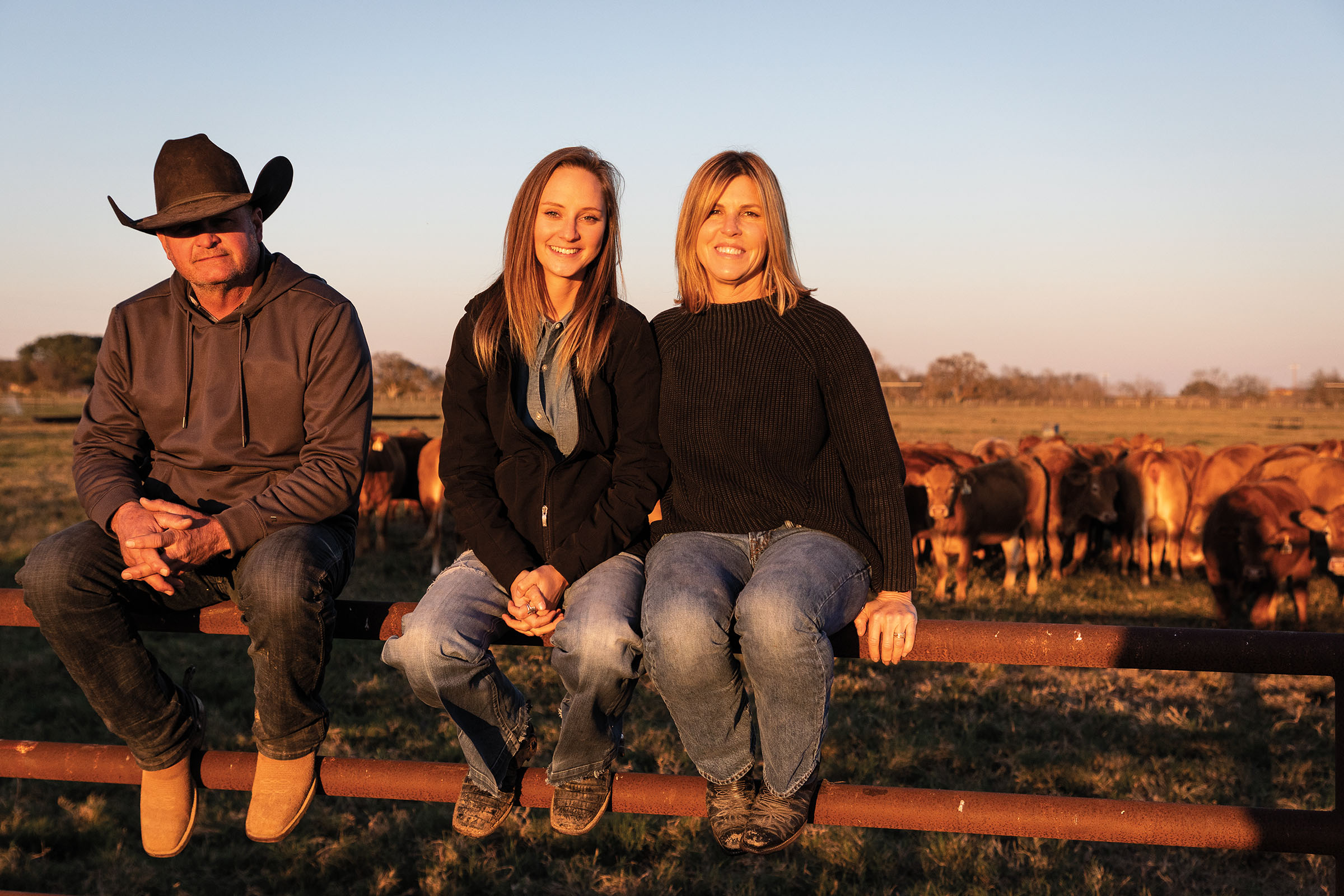 Three people sit on a fence outside of a corral of cows under a blue-pink sky