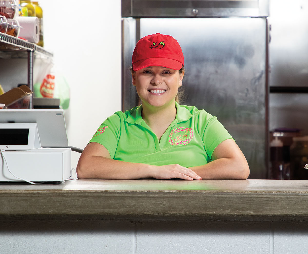 A woman in a green shirt and red hat smiles to the camera in front of a stainless steel kitchen background
