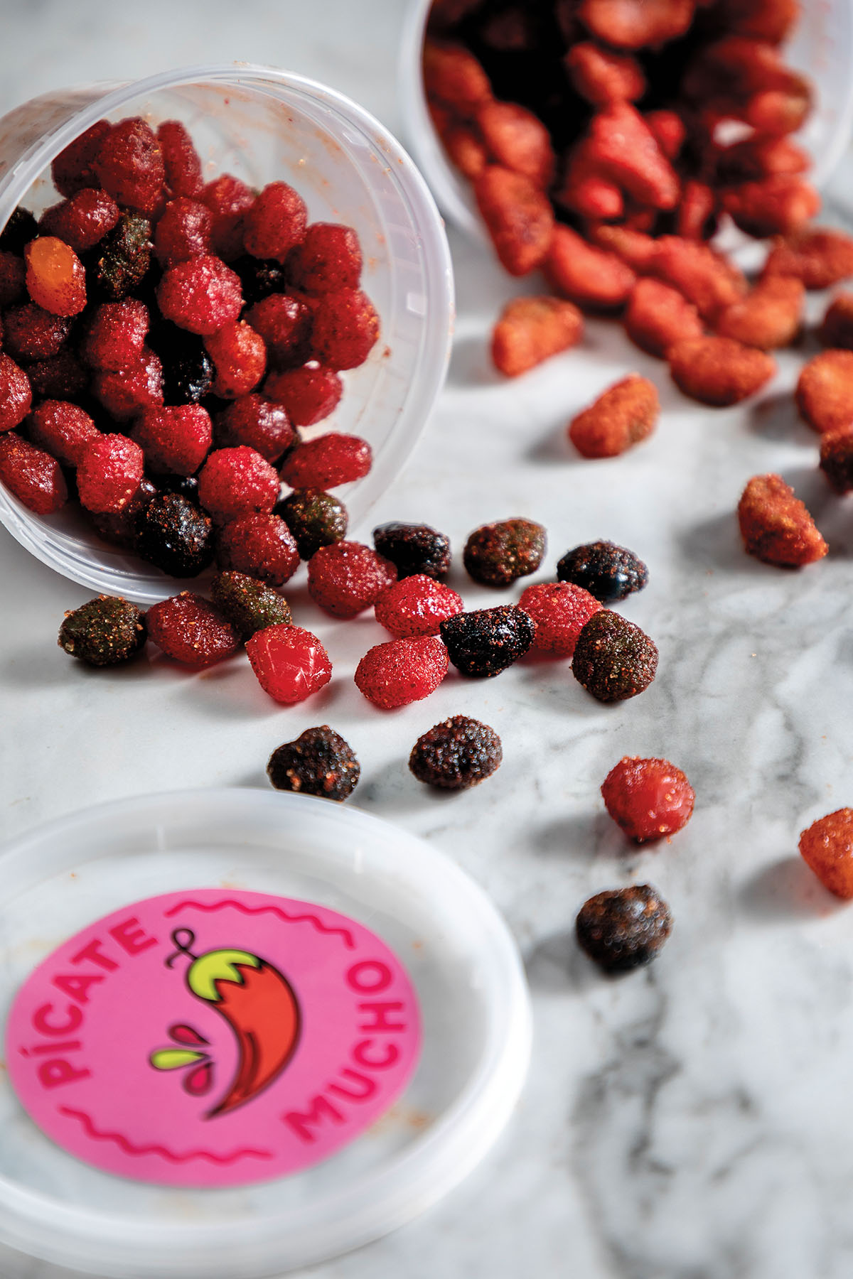 A collection of bright red and purple candies tumble out of a dish with a pink stickered lid reading "Pícate Mucho"