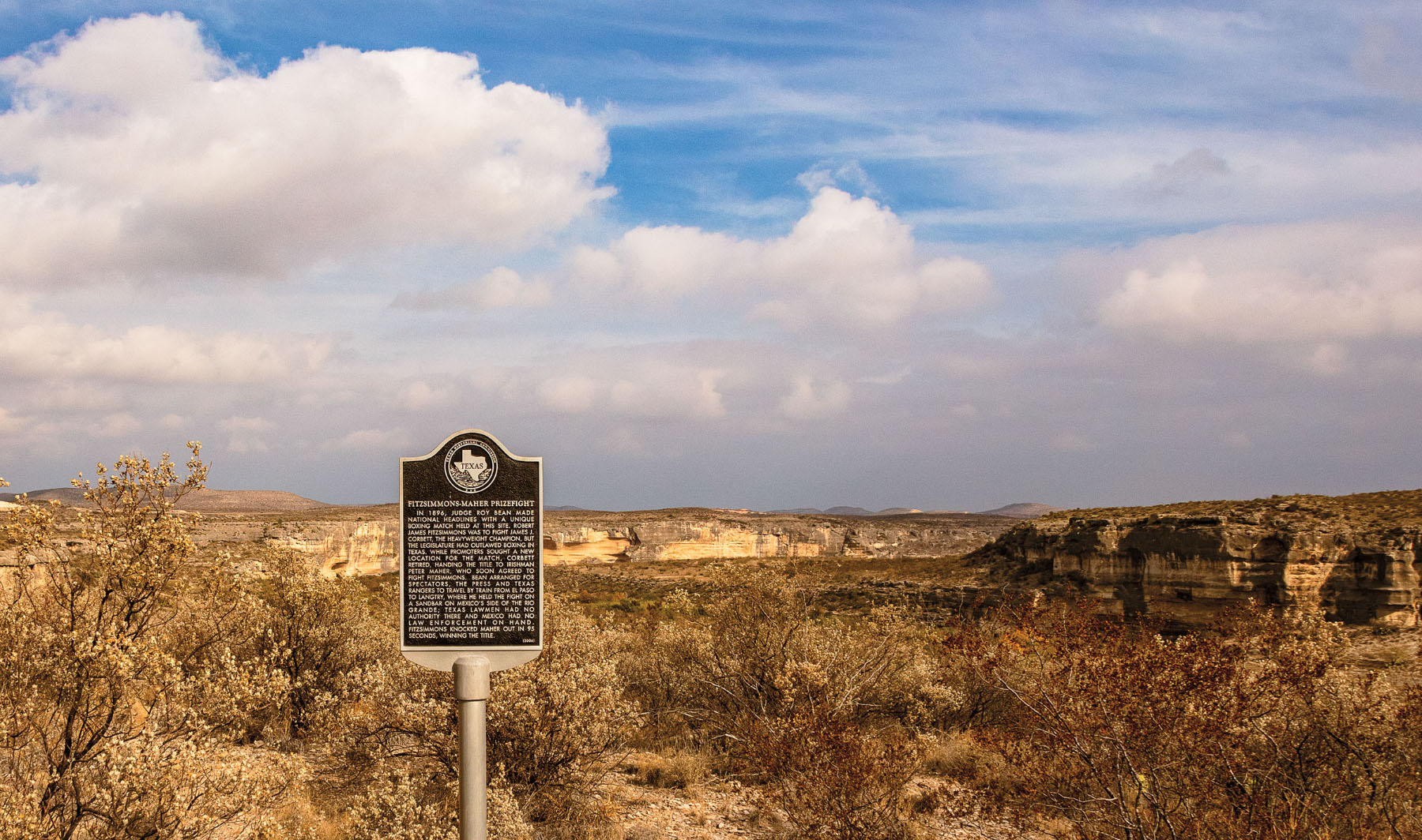 A black and metal Texas historical marker in front of a vast desert landscape under blue sky
