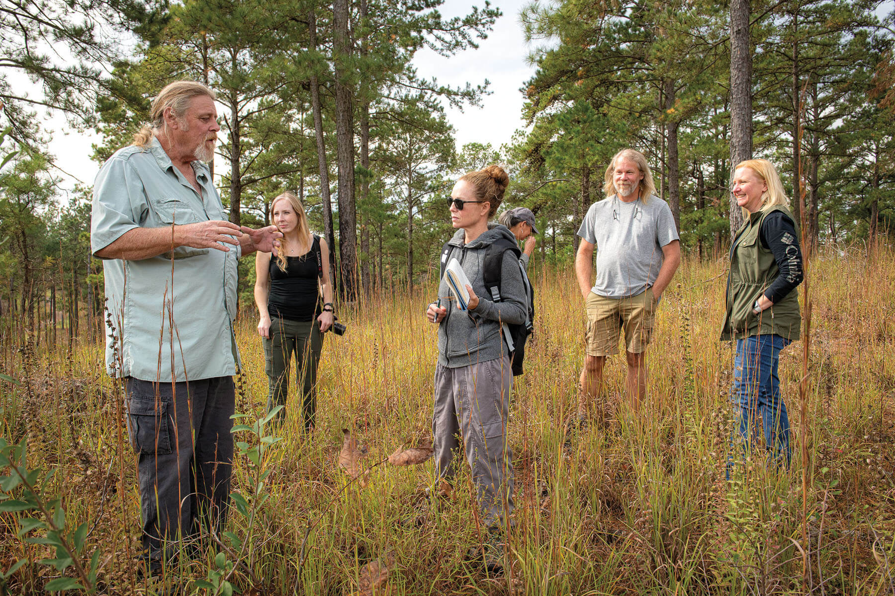 A group of people stand in a field with tall brown grasses beneath green trees