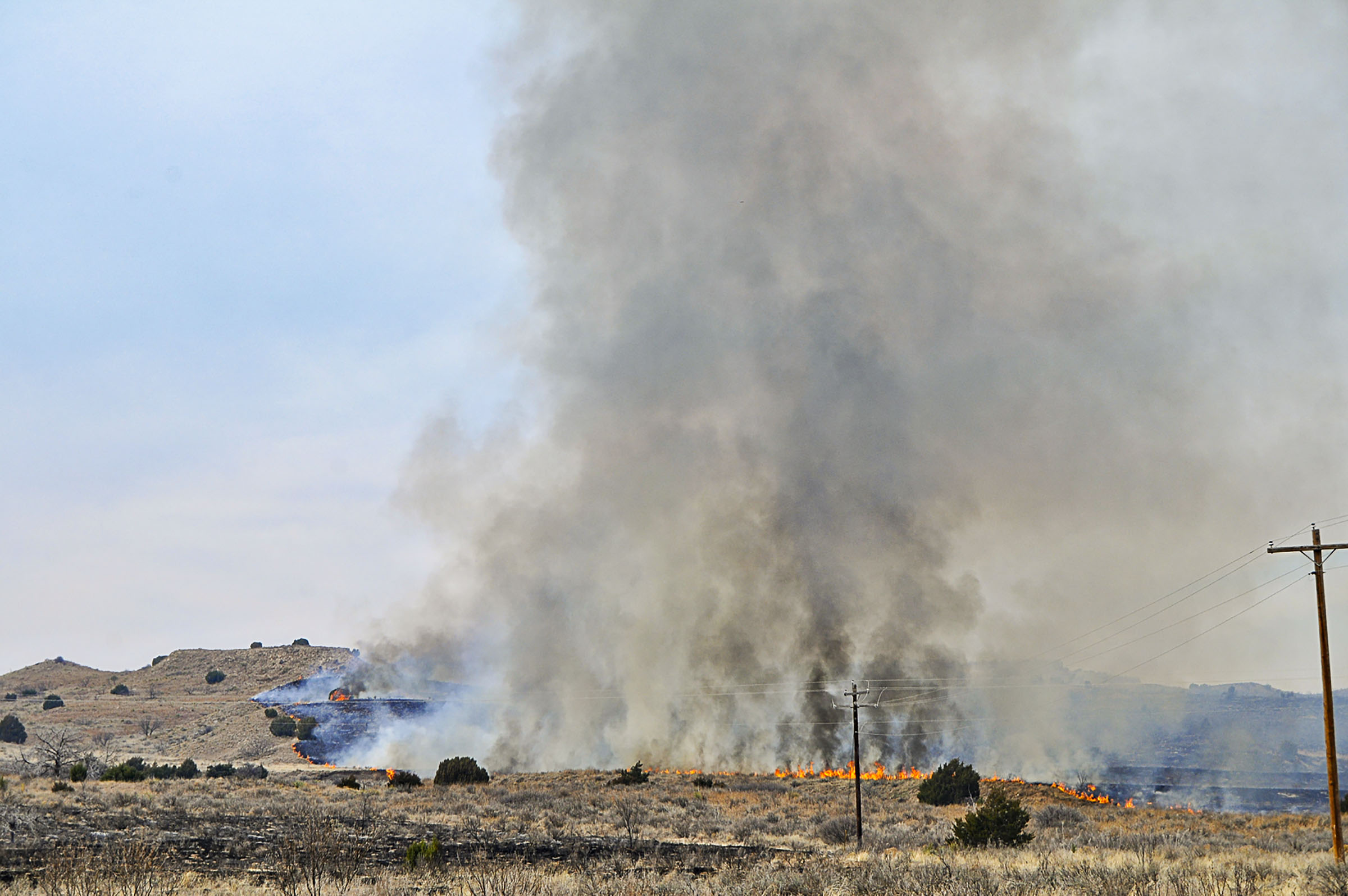A large line of gray smoke rises from a fire along a grassy pasture