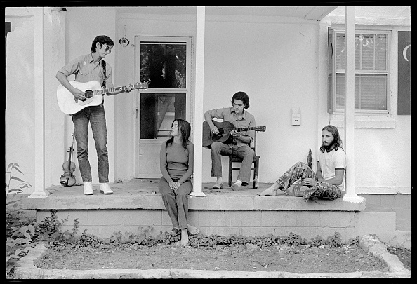 Four people sit on a porch, two playing guitars, in a black and white photograph