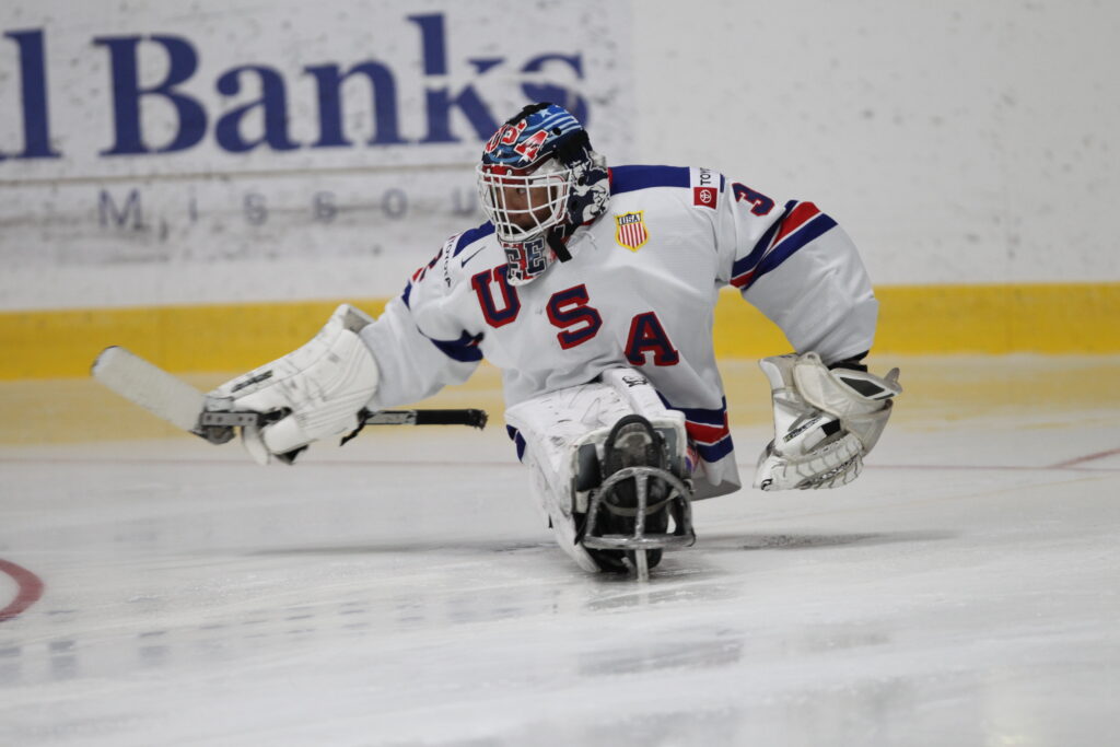 A person in a white jersey reading "USA" participates in sled hockey, wearing a large black face mask and holding a hockey stick