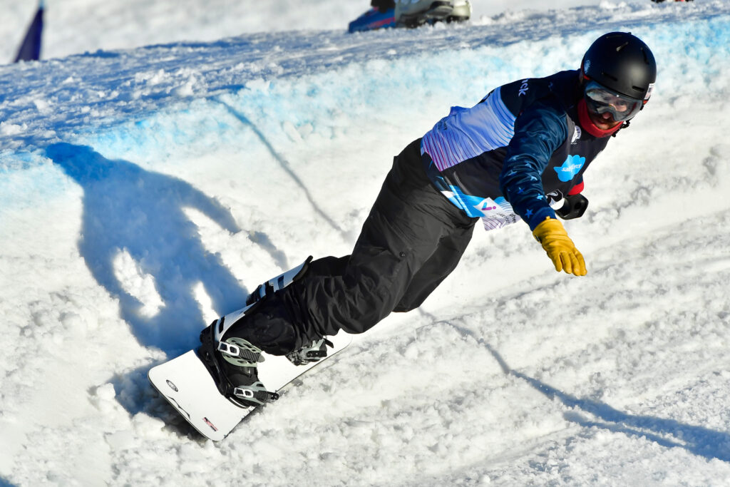A person in a black ski suit and helmet snowboards along a snow-covered ramp with a small amount of blue paint